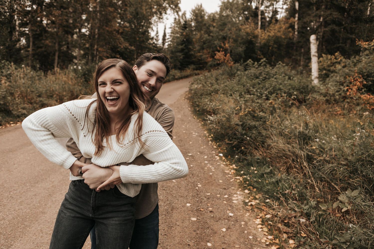Two people share a joyful moment together while embracing on a dirt path surrounded by autumn foliage.