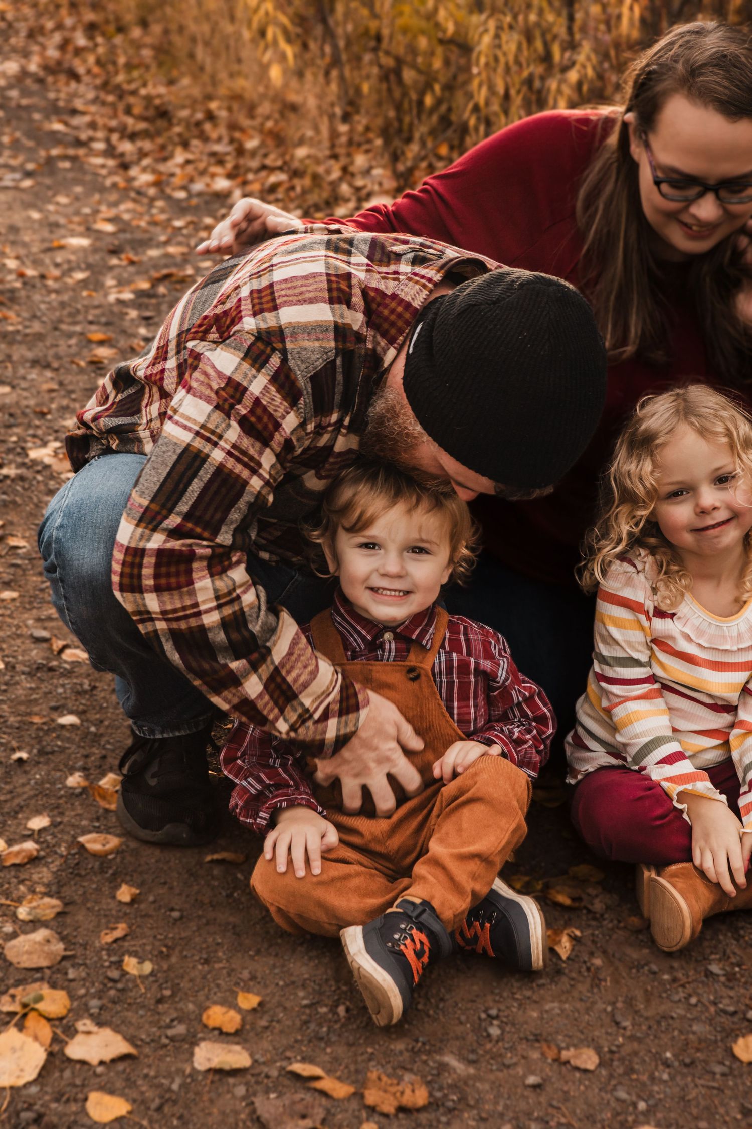 Family sits together on autumn ground with fallen leaves in warm earth tones and plaid clothing.
