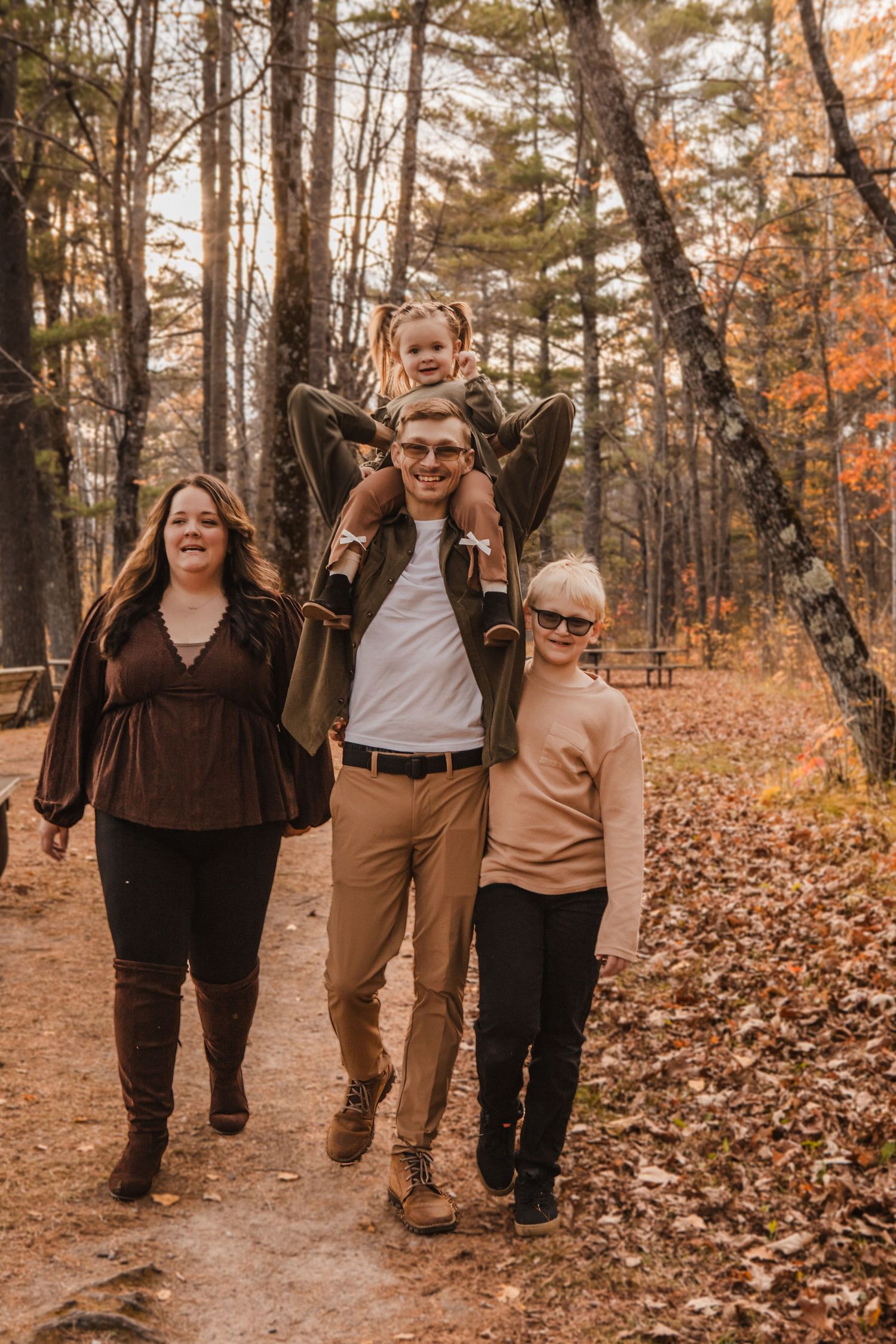 A group walks together on a wooded trail surrounded by autumn foliage and fallen leaves on a sunny day.
