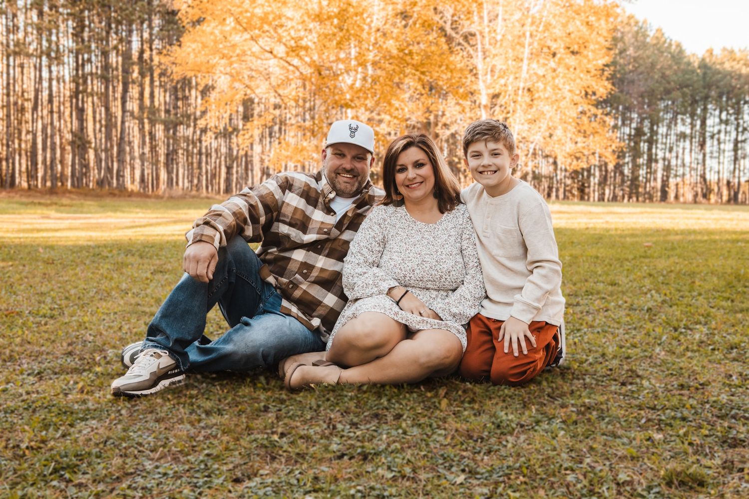 A family sits together on autumn grass with golden trees in the background during a fall photo session.