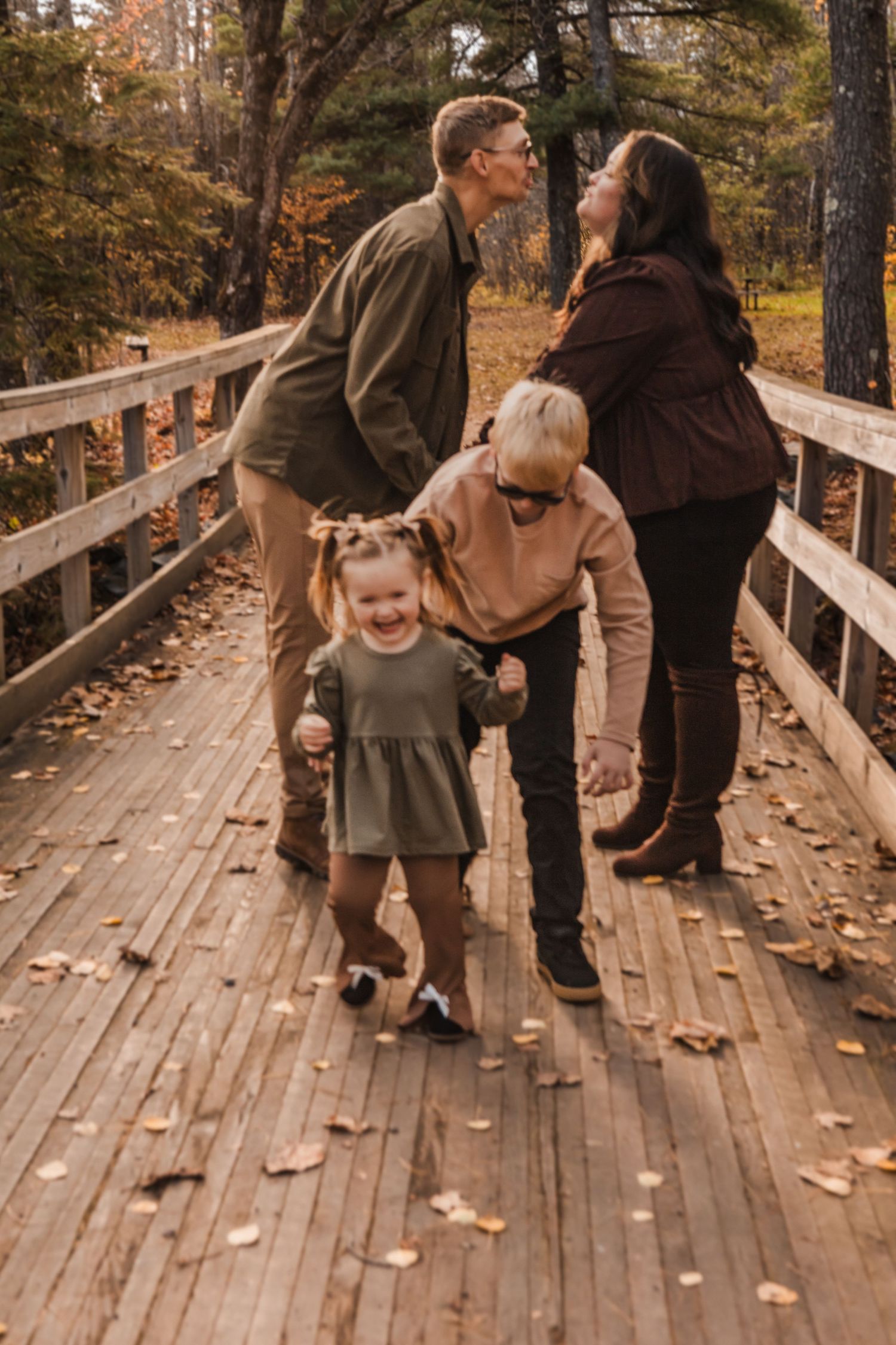 Family walking together on a wooden boardwalk surrounded by autumn trees with fallen leaves.