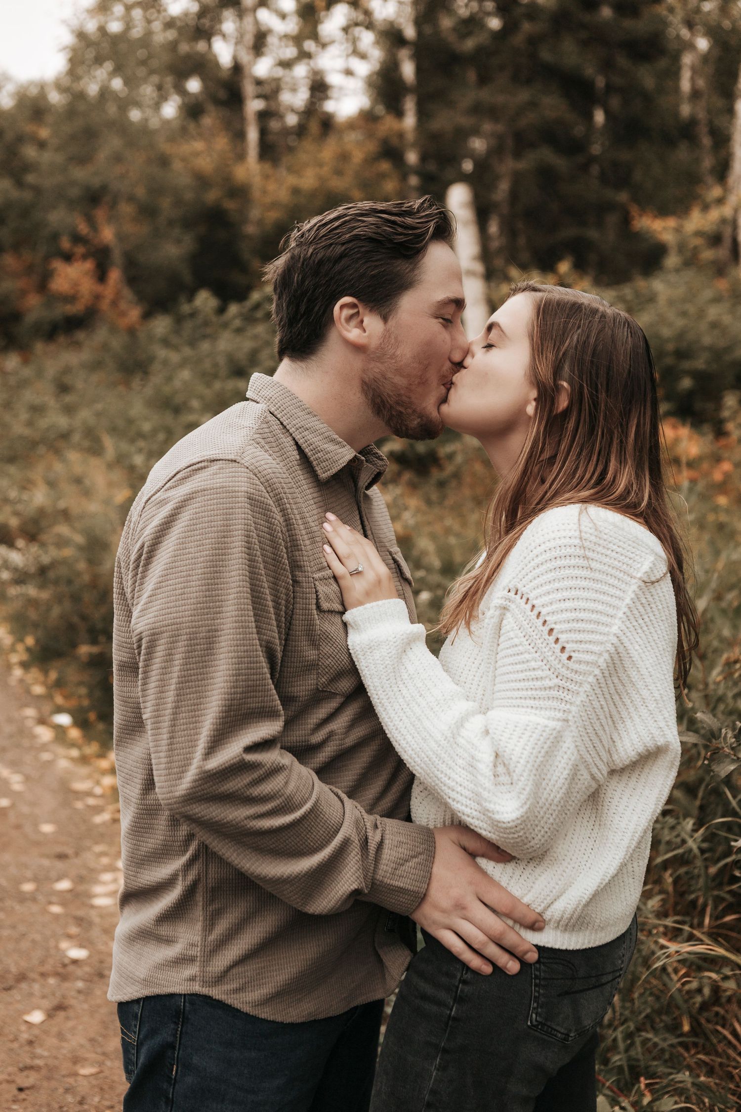 A romantic autumn scene of a couple sharing a tender kiss on a wooded path while wearing cozy sweaters.