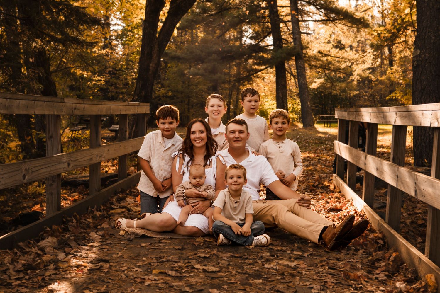 A family wearing white and khaki outfits poses together in a golden autumn forest setting during sunset.