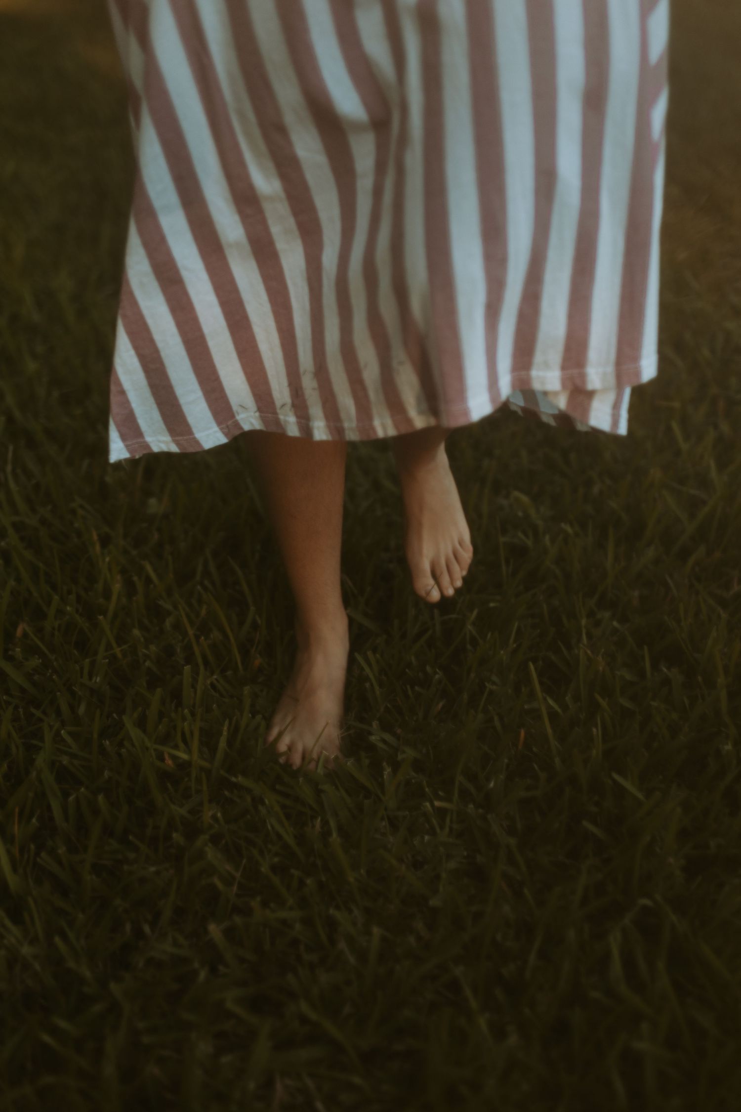 Barefoot person wearing a striped dress standing in dark grass during dusk or twilight.