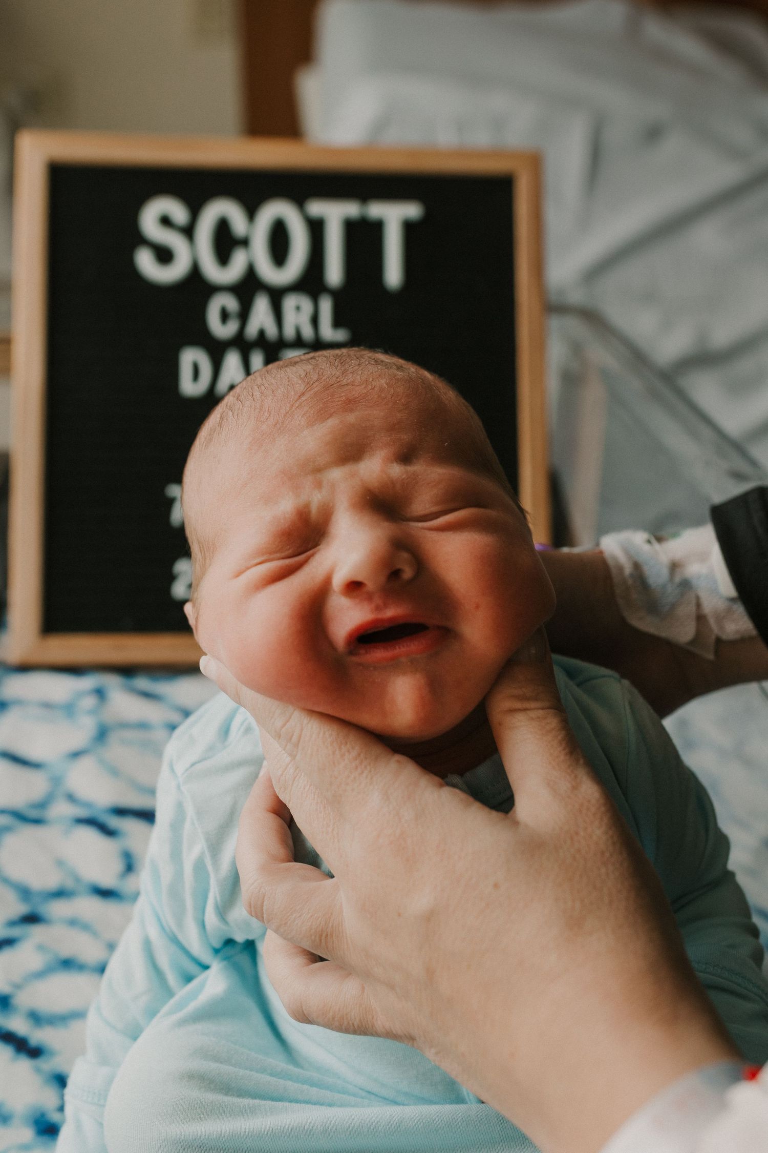A newborn baby in light blue clothing smiles while being held, with a name board visible in the background.