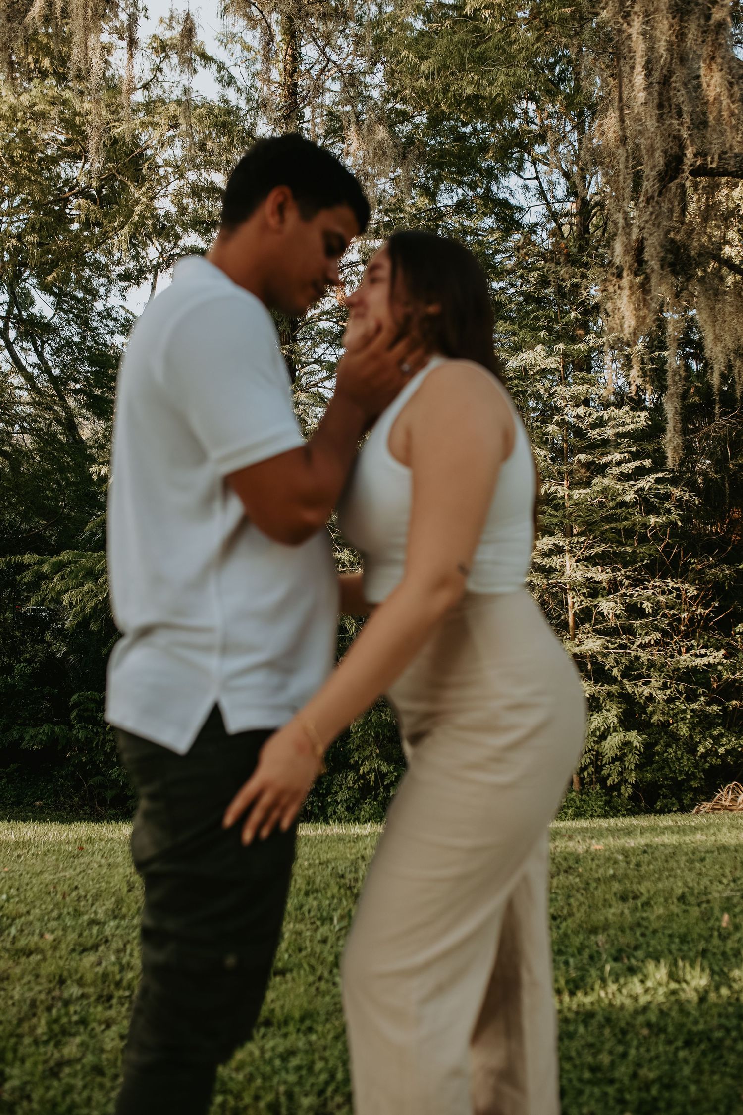 A romantic couple in white outfits share an intimate moment outdoors with Spanish moss hanging from trees in the background.