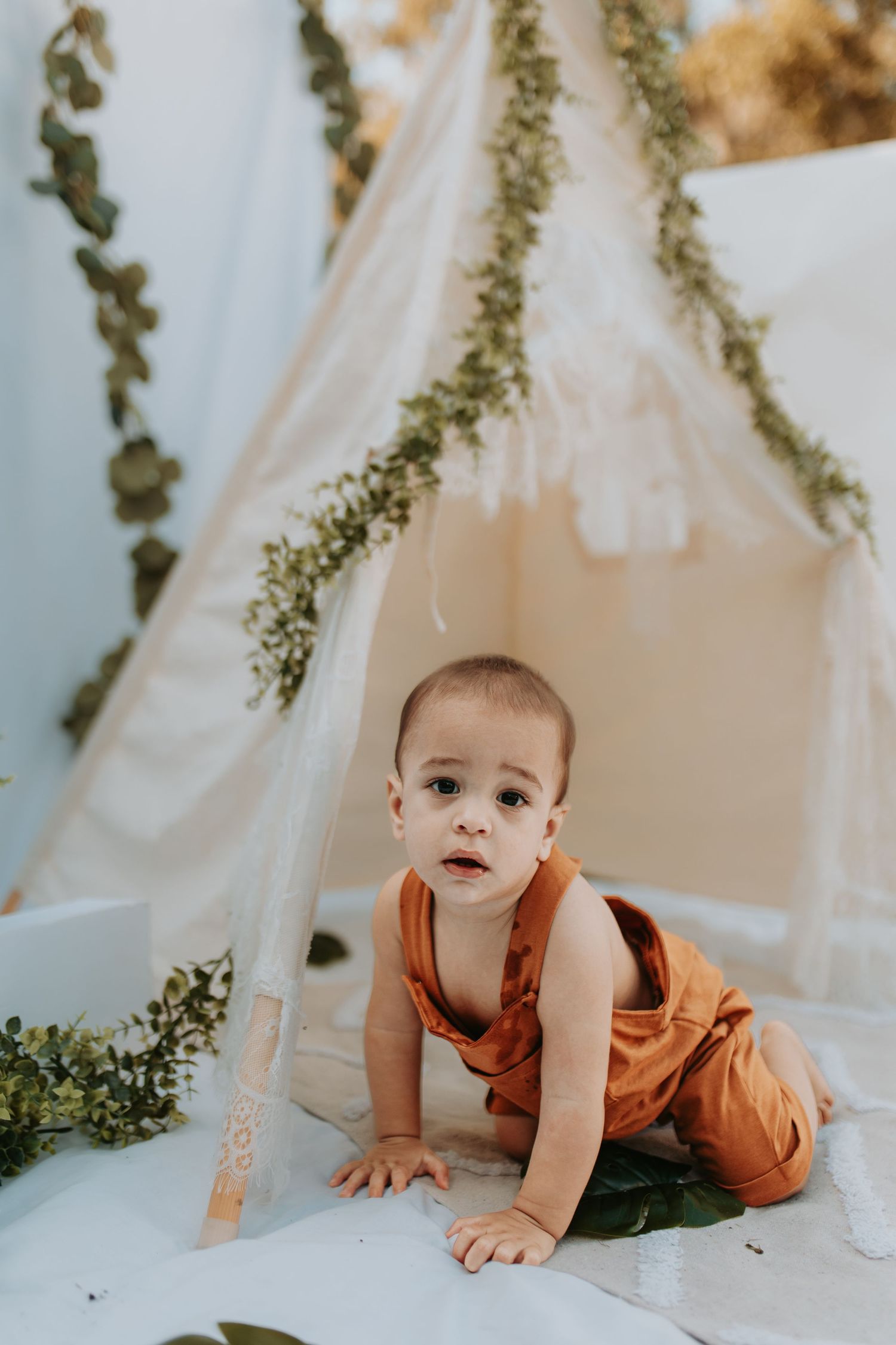 Baby in orange overalls crawls inside dreamy tent setting with hanging greenery garlands and white fabric backdrop.