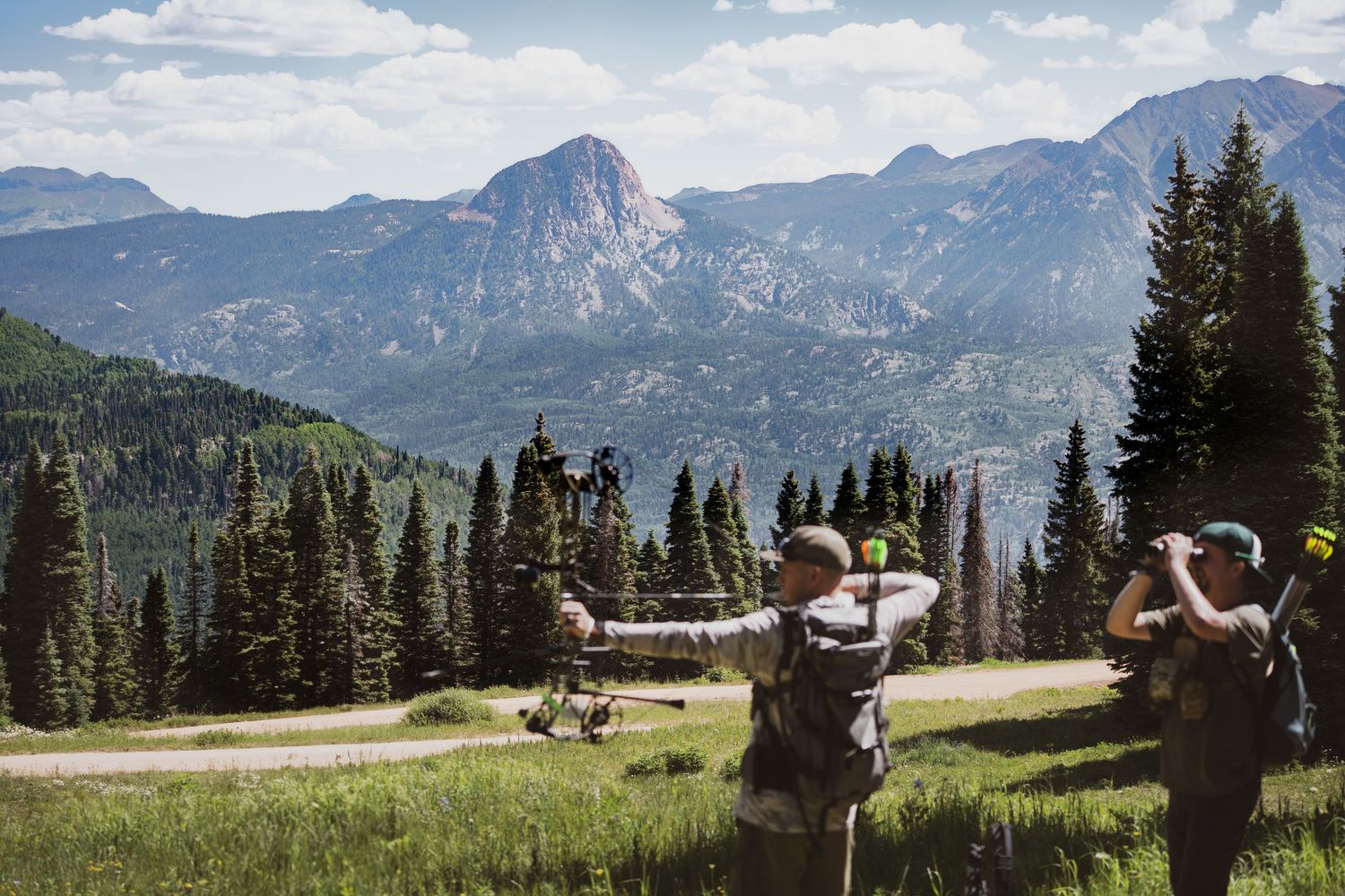 Landscape shot of mountains with two men in front with a bow and binoculars