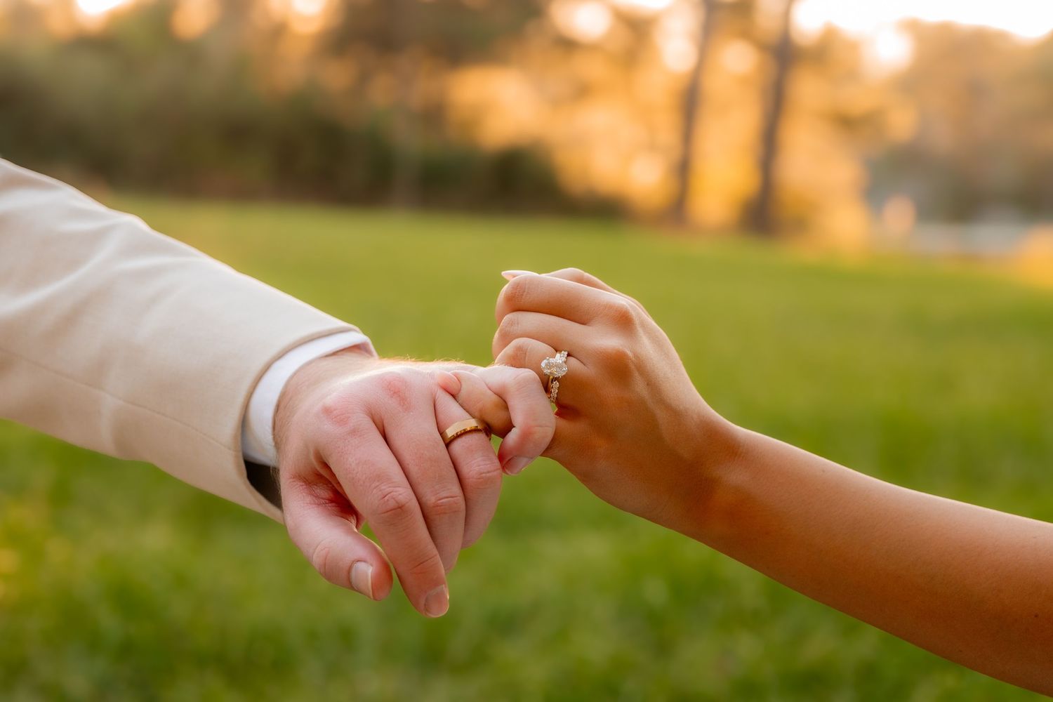 Newlyweds hold hands showing wedding rings at sunset in a lush green outdoor setting.