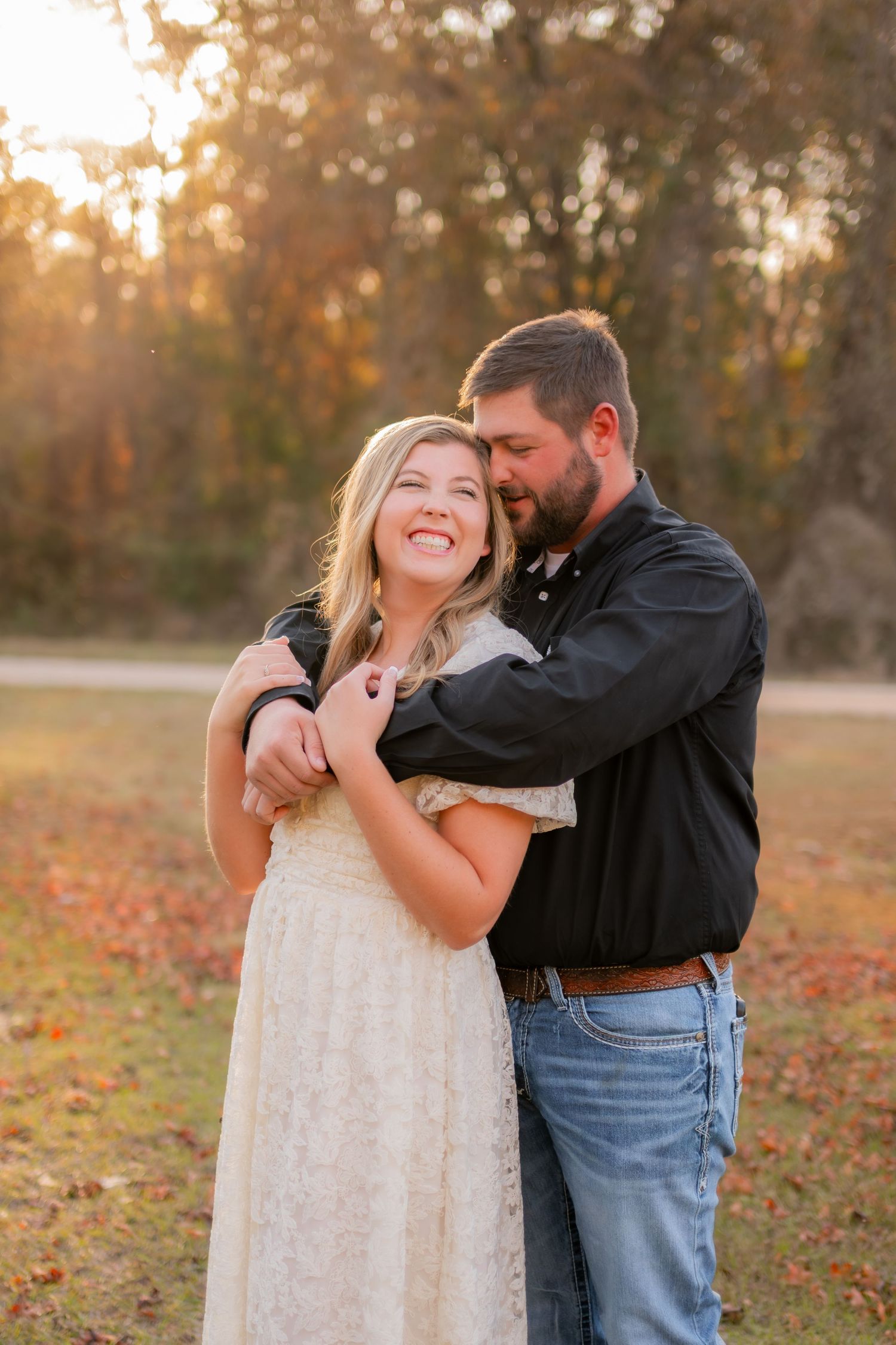 Happy couple embraces at golden hour in an autumn outdoor setting, woman in cream dress smiling joyfully.