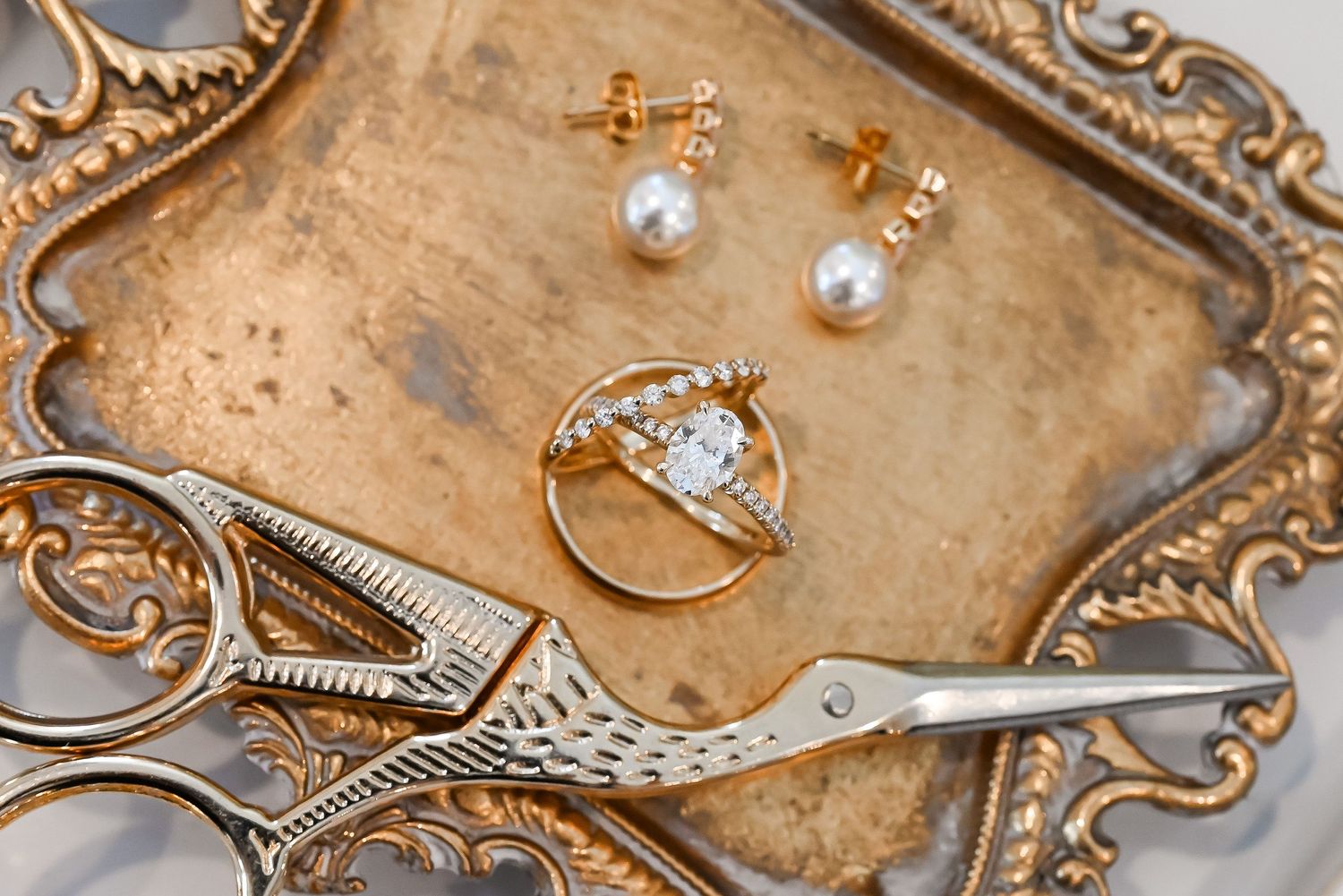 Diamond engagement ring with halo setting beside pearl drop earrings on ornate gold tray.