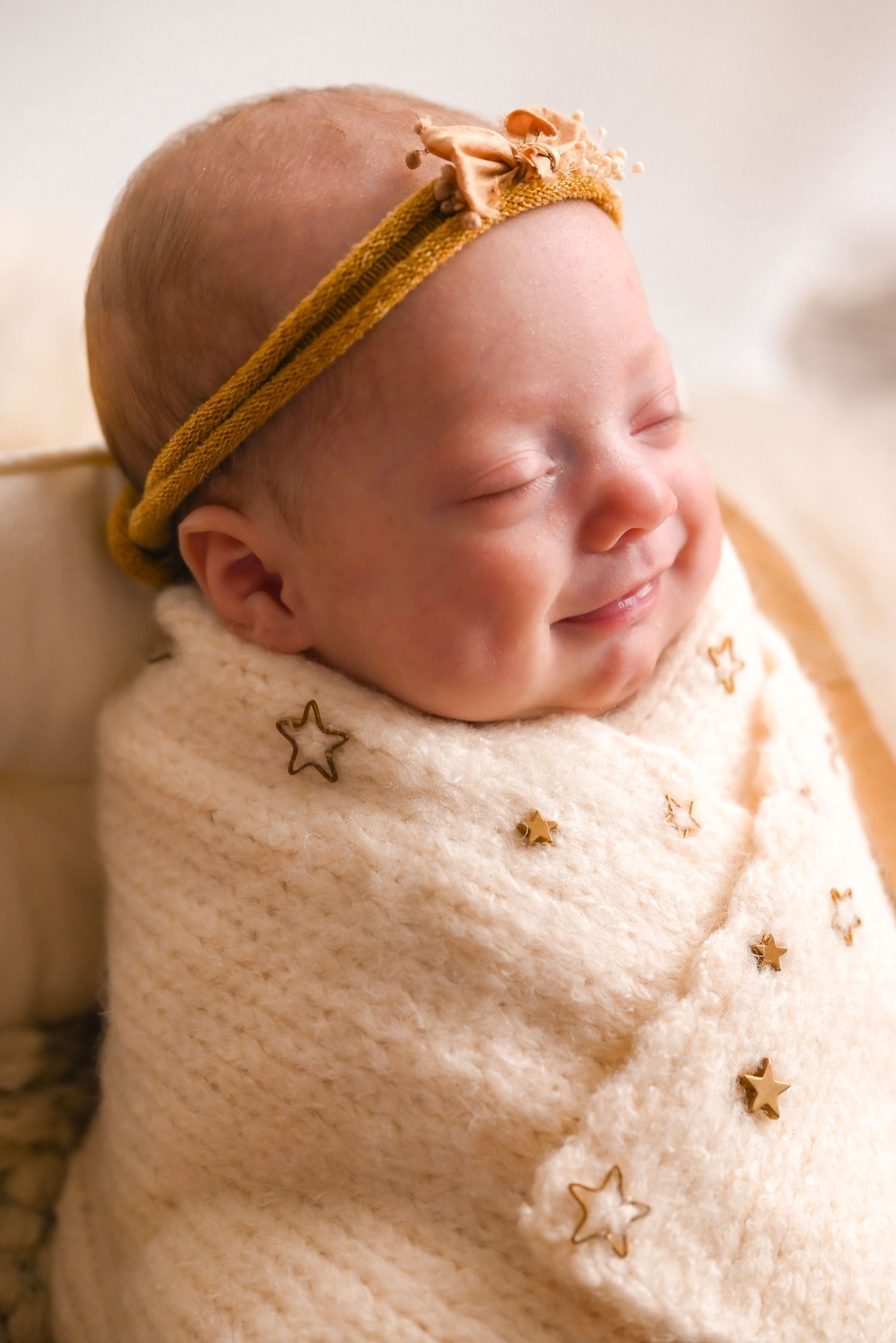 Smiling newborn baby wrapped in cream star-print swaddle wearing a mustard yellow bow headband.