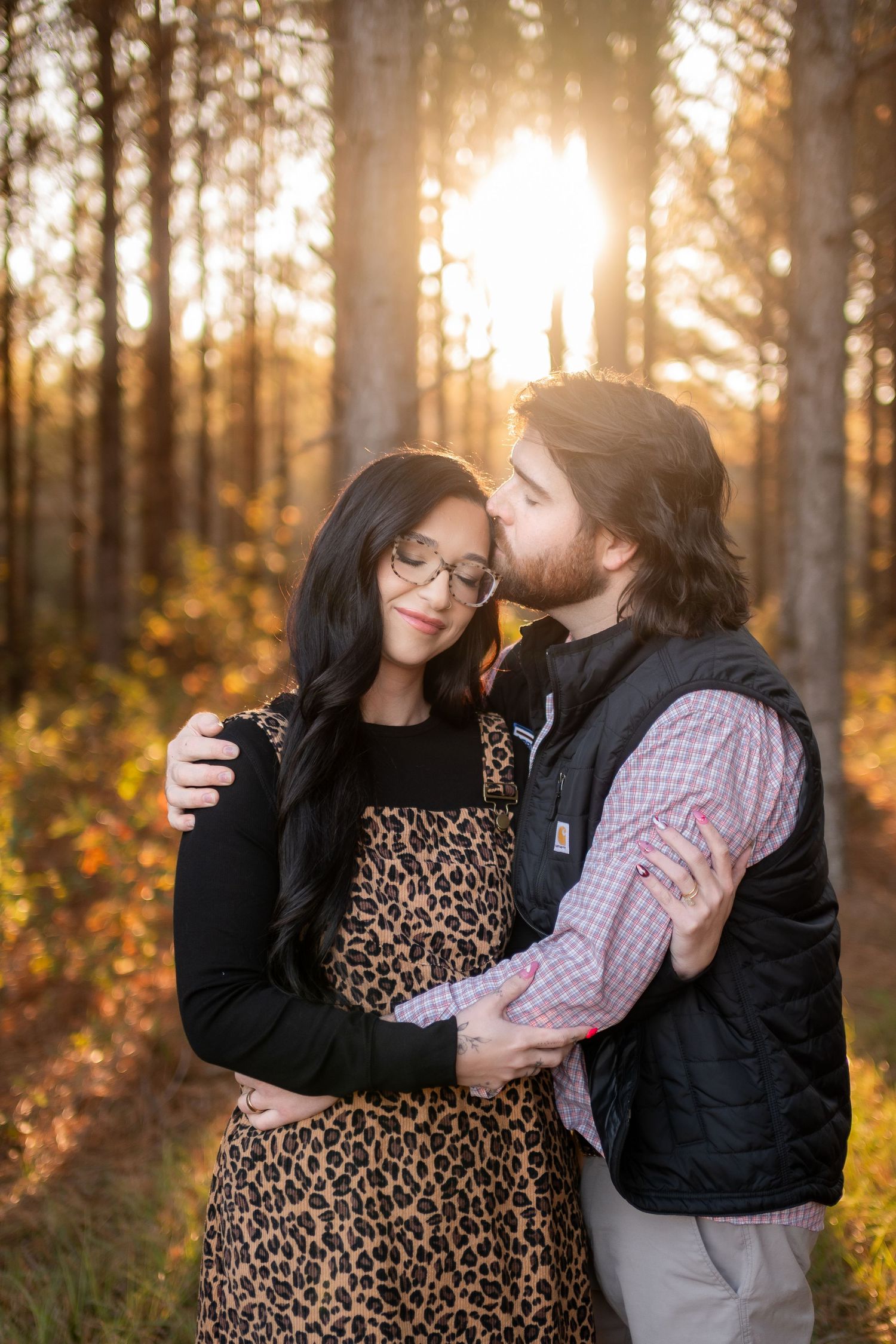 Man kissing woman on the forehead in a sun-drenched forest during golden hour engagement session.