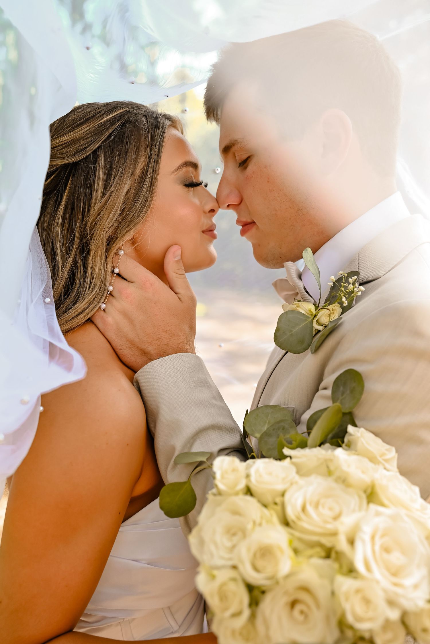 Bride and groom share an intimate moment under a veil, nearly kissing, with a bouquet of white roses.