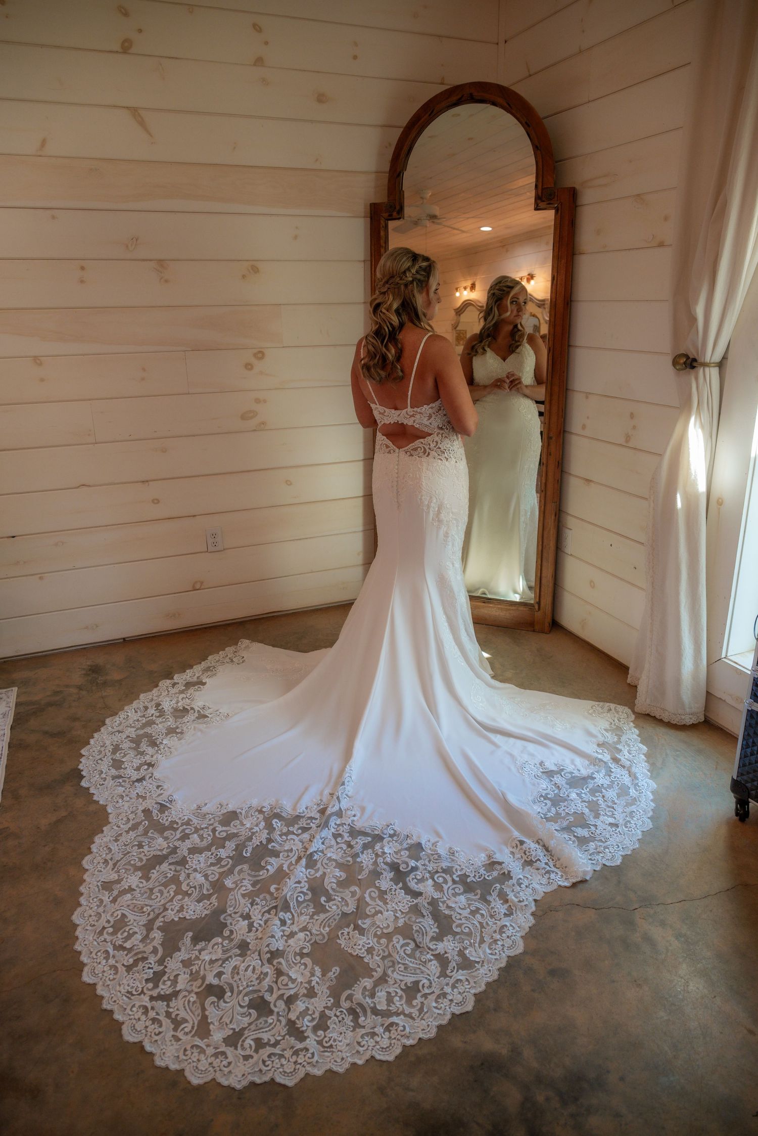 Bride in fitted lace train wedding gown viewed from behind reflecting in ornate mirror in rustic white wood room.
