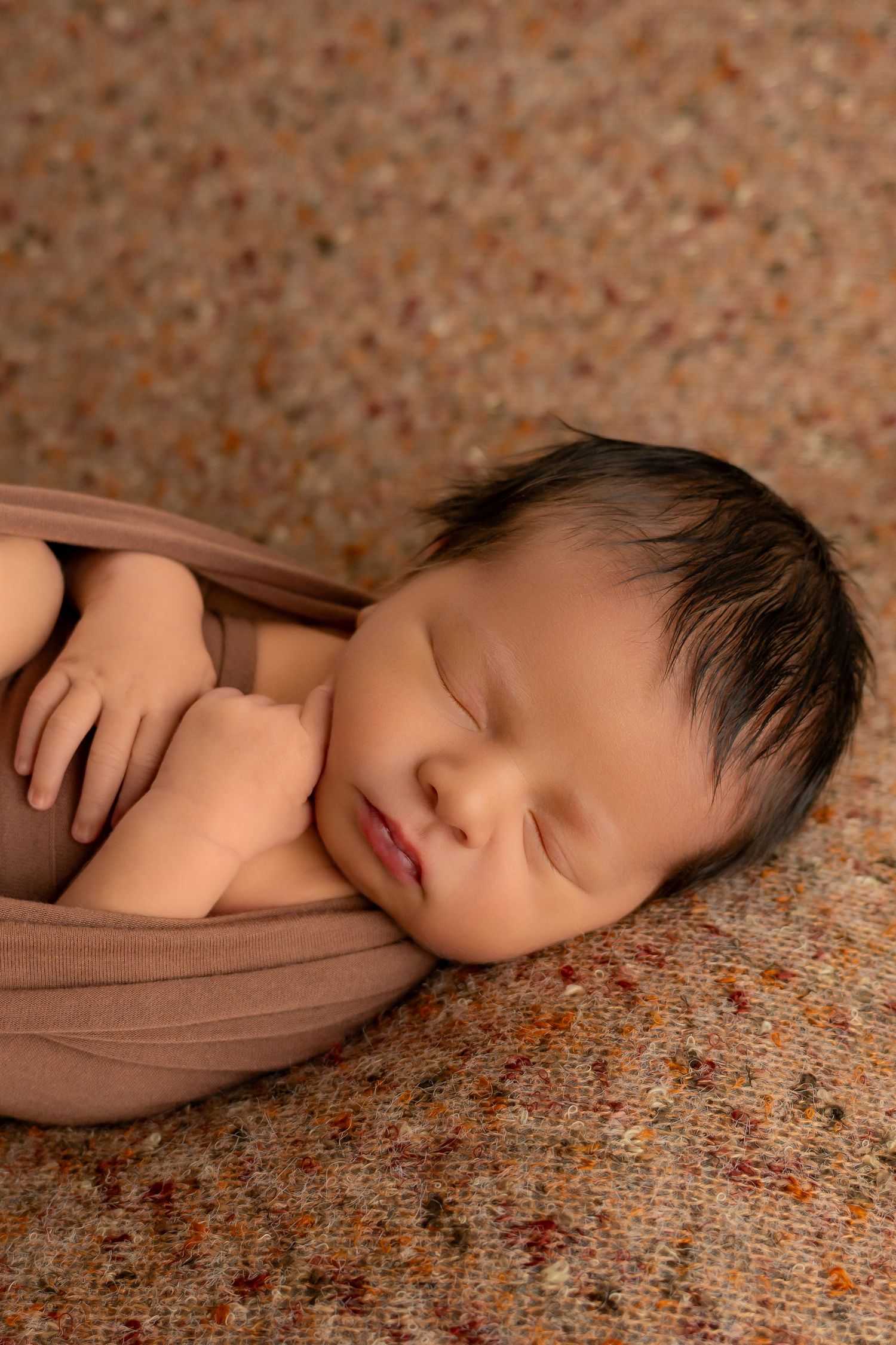 Peaceful newborn baby with dark hair wrapped in brown swaddle sleeping on textured speckled backdrop.