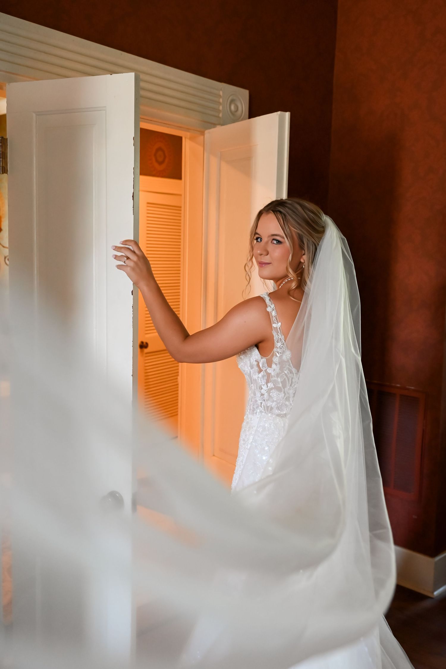 Elegant bride in lace wedding gown and veil opening double doors in warmly lit room.