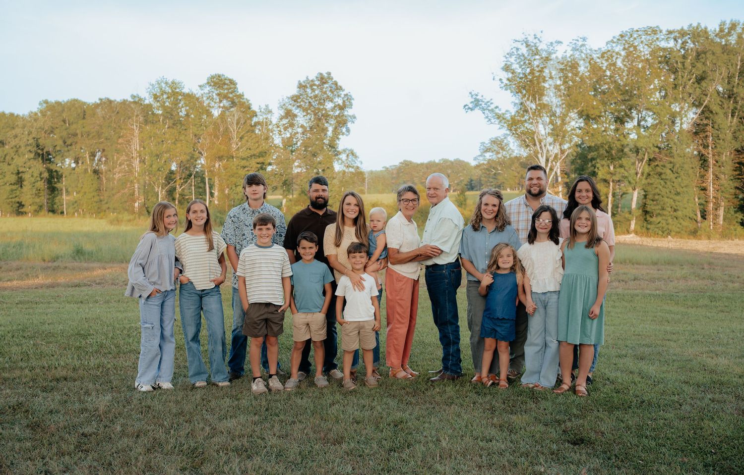 Large multigenerational family posing together outdoors in a green field surrounded by tall trees during golden hour.