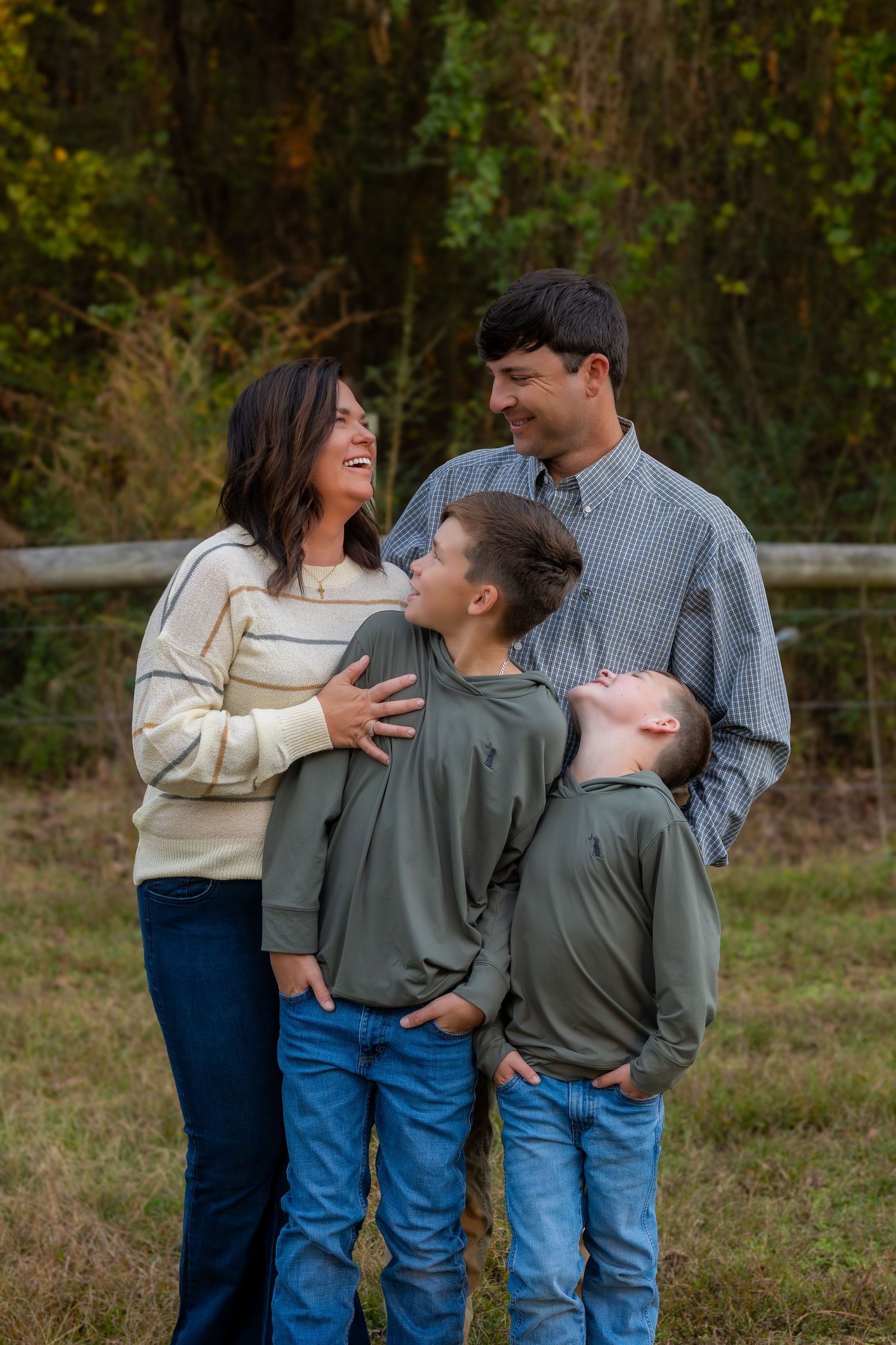 Happy family of four with two boys laughing together outdoors near rustic wooden fence in autumn setting.