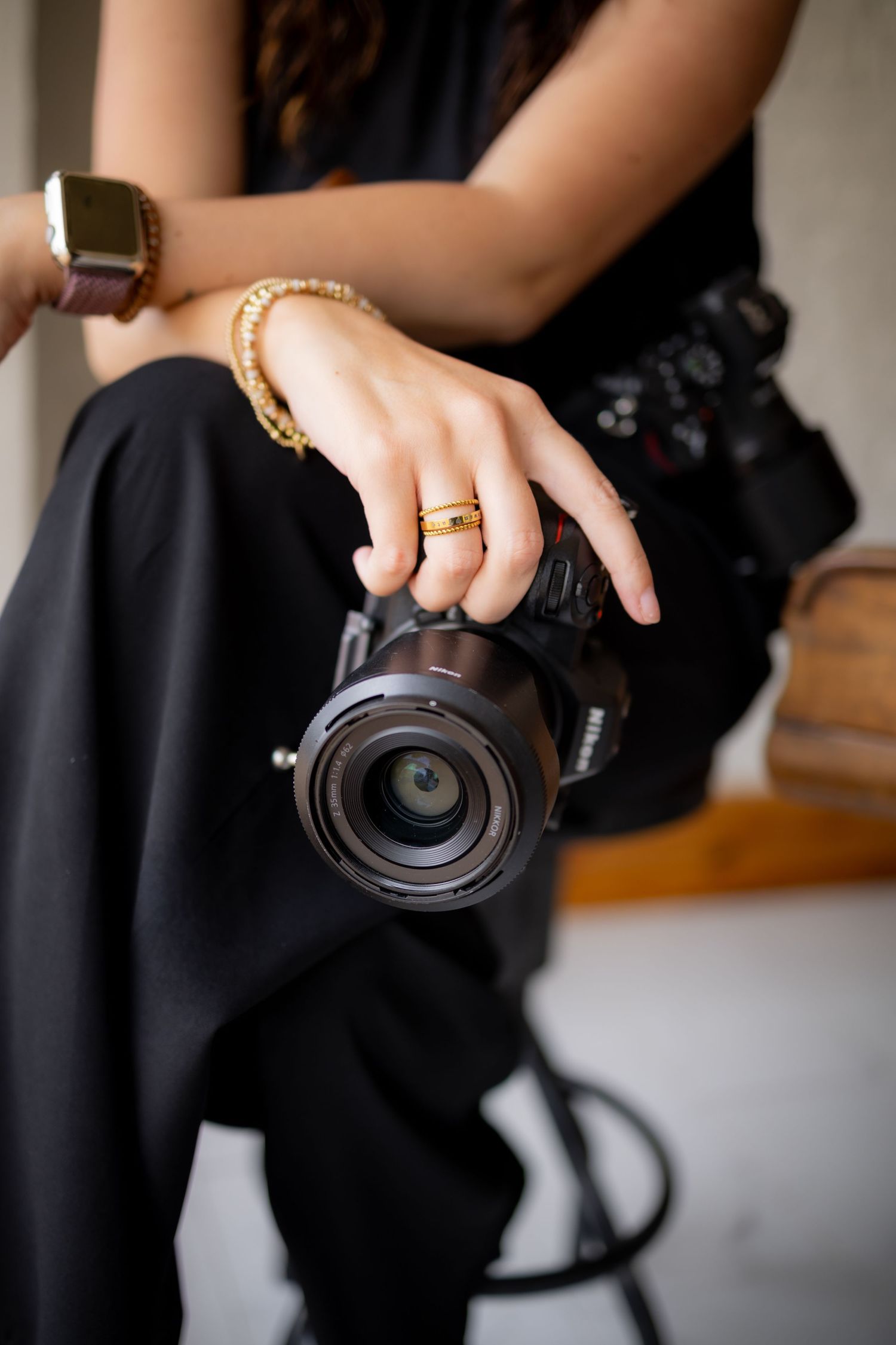Woman in black outfit holding a professional camera, wearing gold rings and bracelets, seated on a stool.