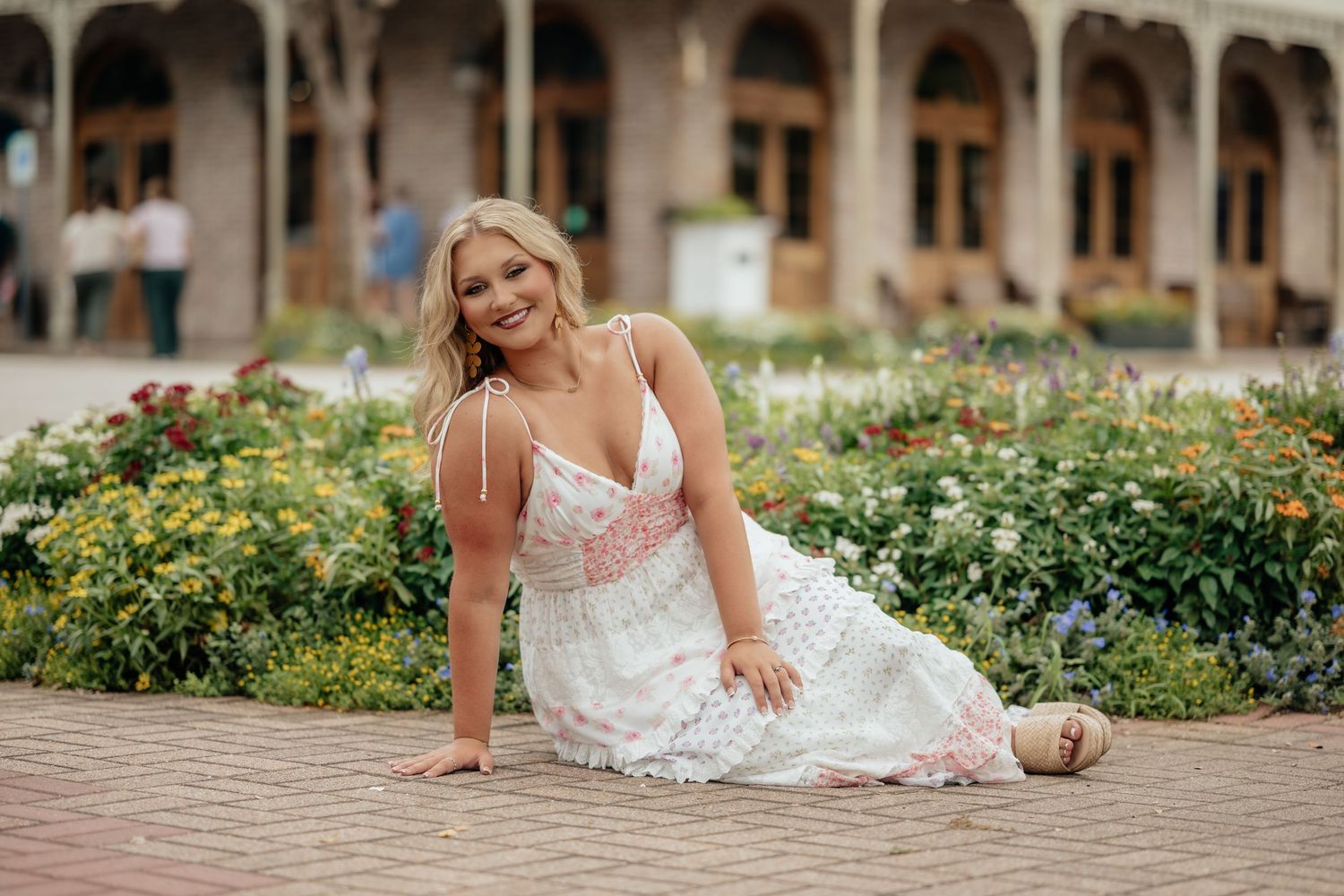 Woman in white floral sundress sitting gracefully on brick path beside colorful garden blooms near elegant building.