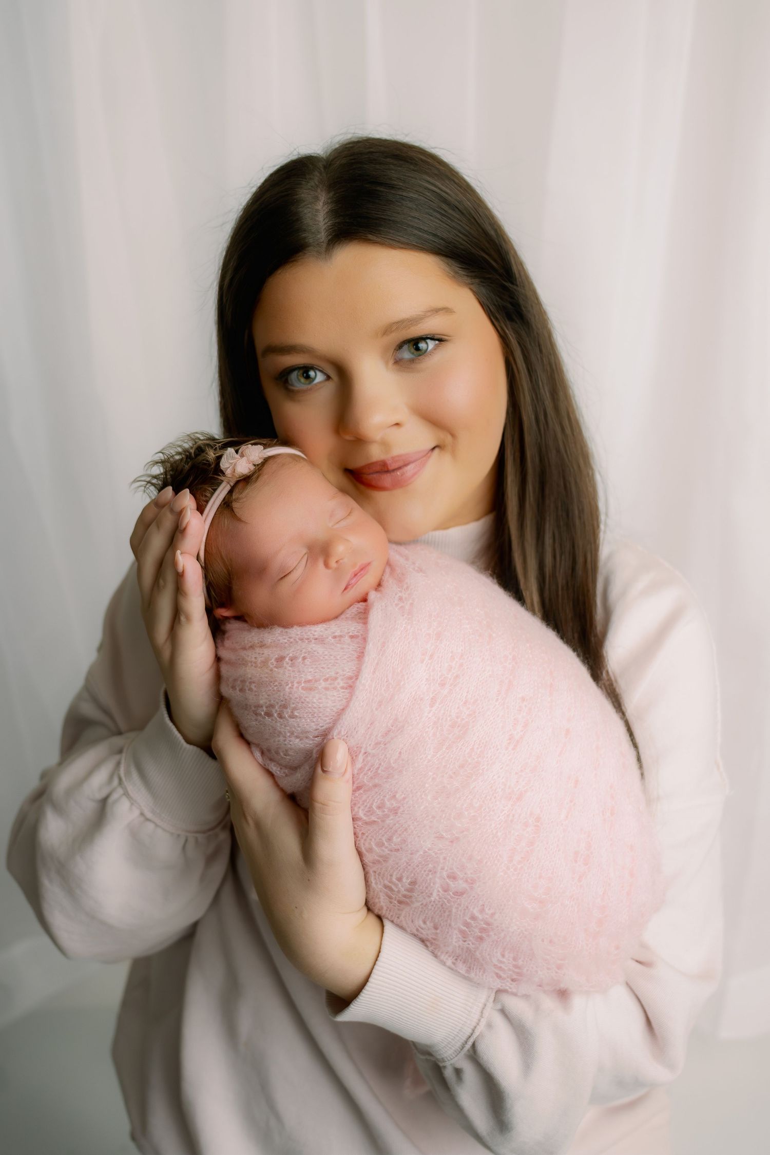 Young mother in white top tenderly holding sleeping newborn baby wrapped in pink lace blanket.