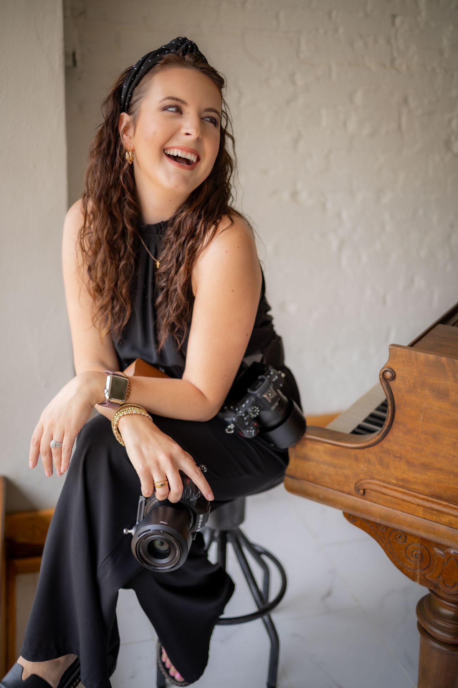 Laughing woman in black outfit sitting on stool holding camera near vintage piano in white brick room.