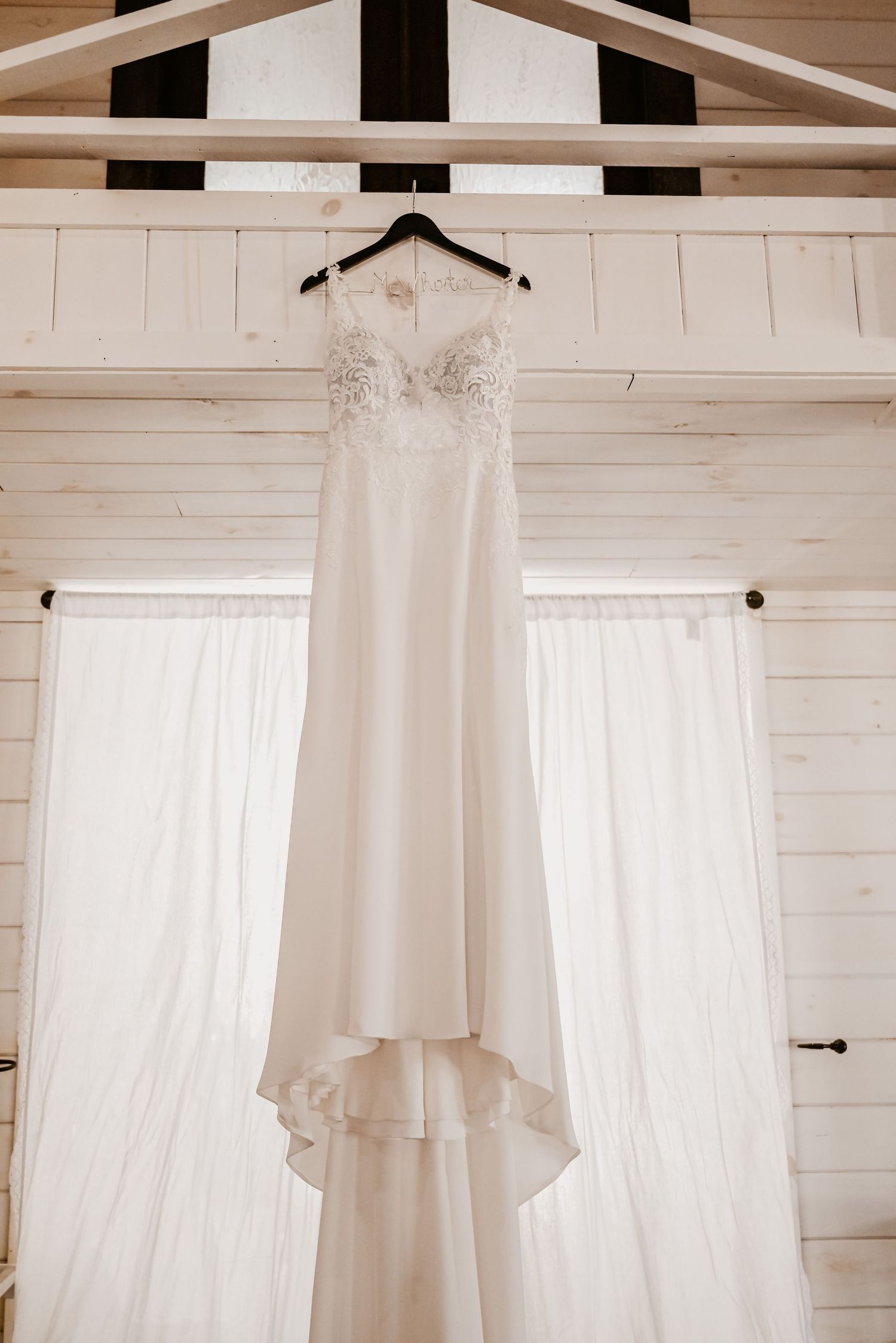 Elegant white lace wedding gown hanging on a hanger in a bright white wood-paneled bridal suite.
