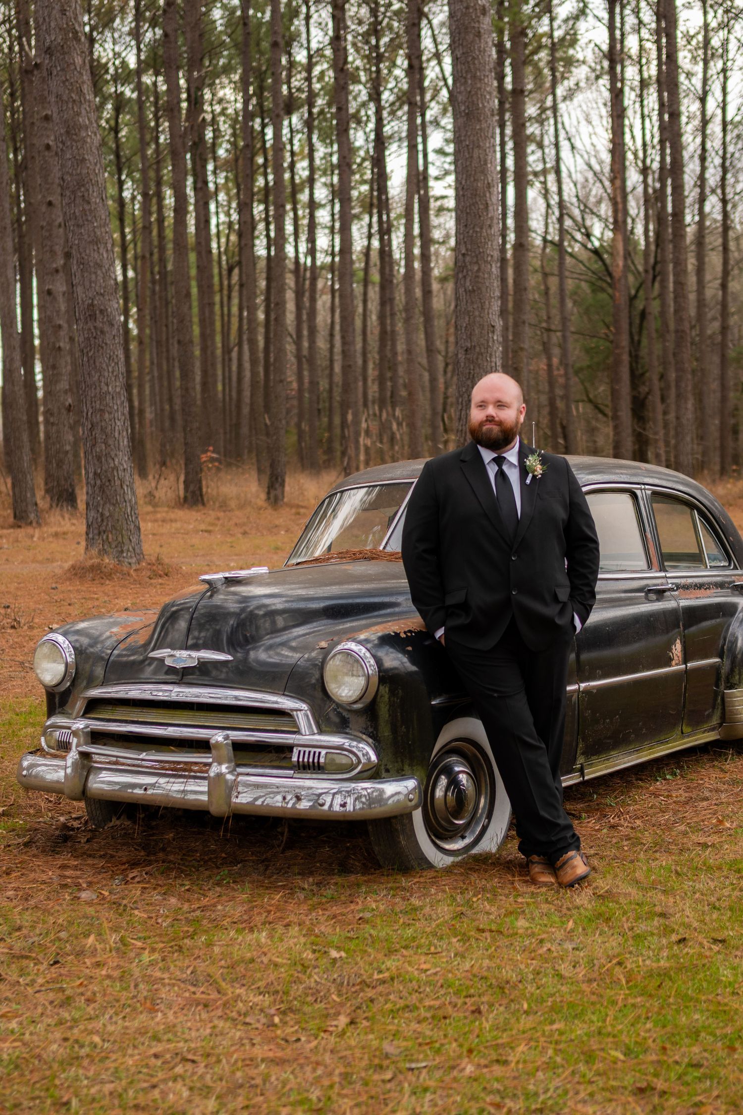 Groom in black suit leaning against vintage classic black car in pine forest setting.