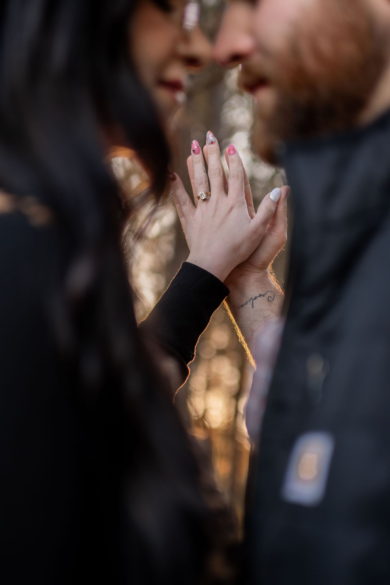 Close-up of couple holding hands showing engagement ring with bokeh forest background.