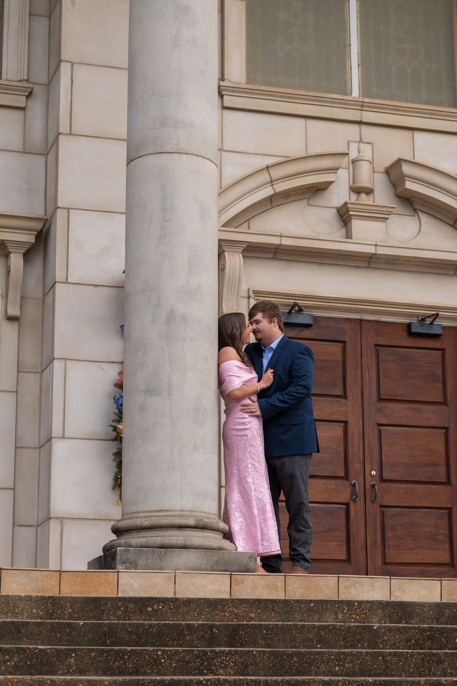 Couple sharing a romantic moment on the steps of a grand stone building with tall columns and wooden doors.