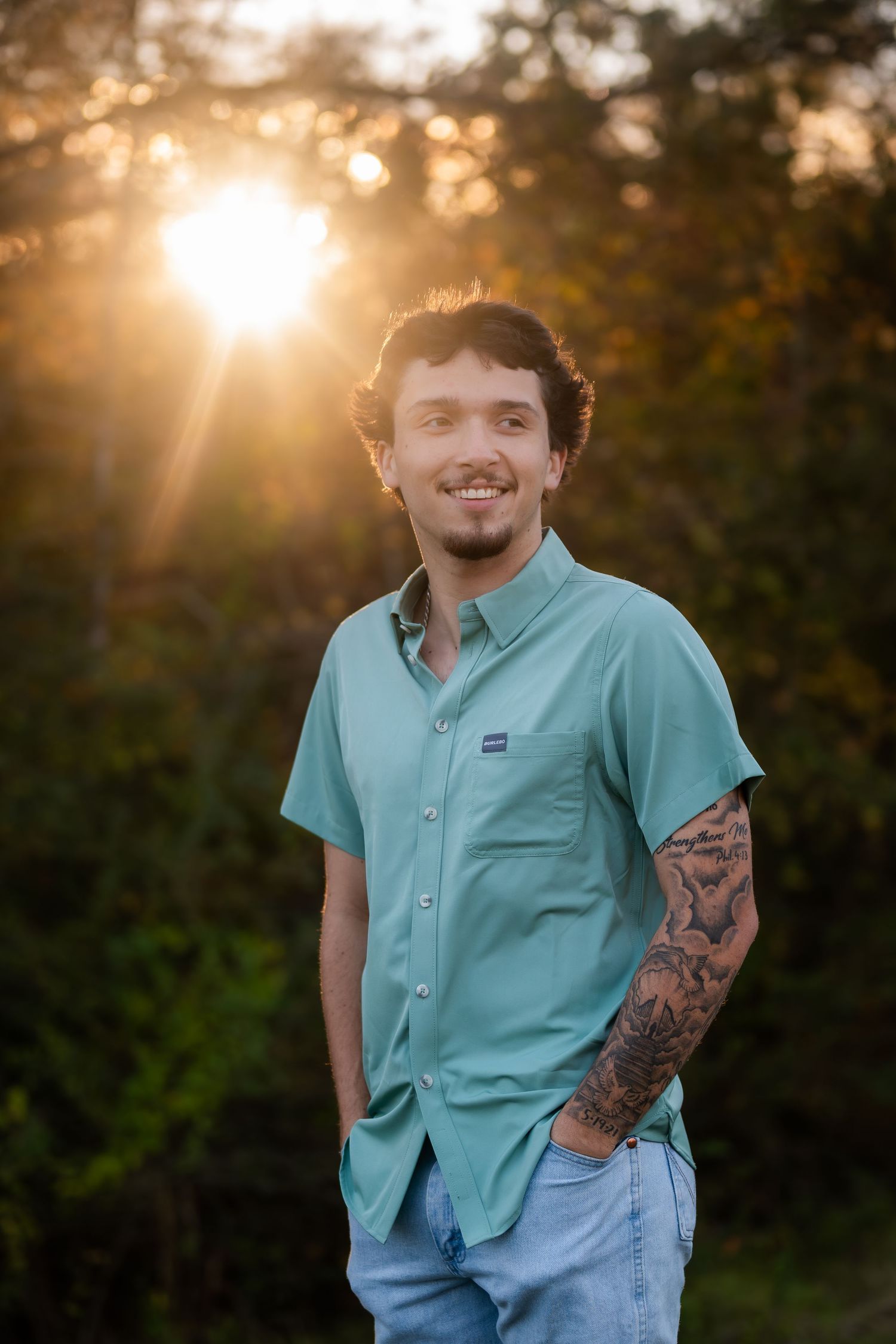 Smiling young man with tattoo sleeve wearing teal shirt standing outdoors during golden sunset backlight.