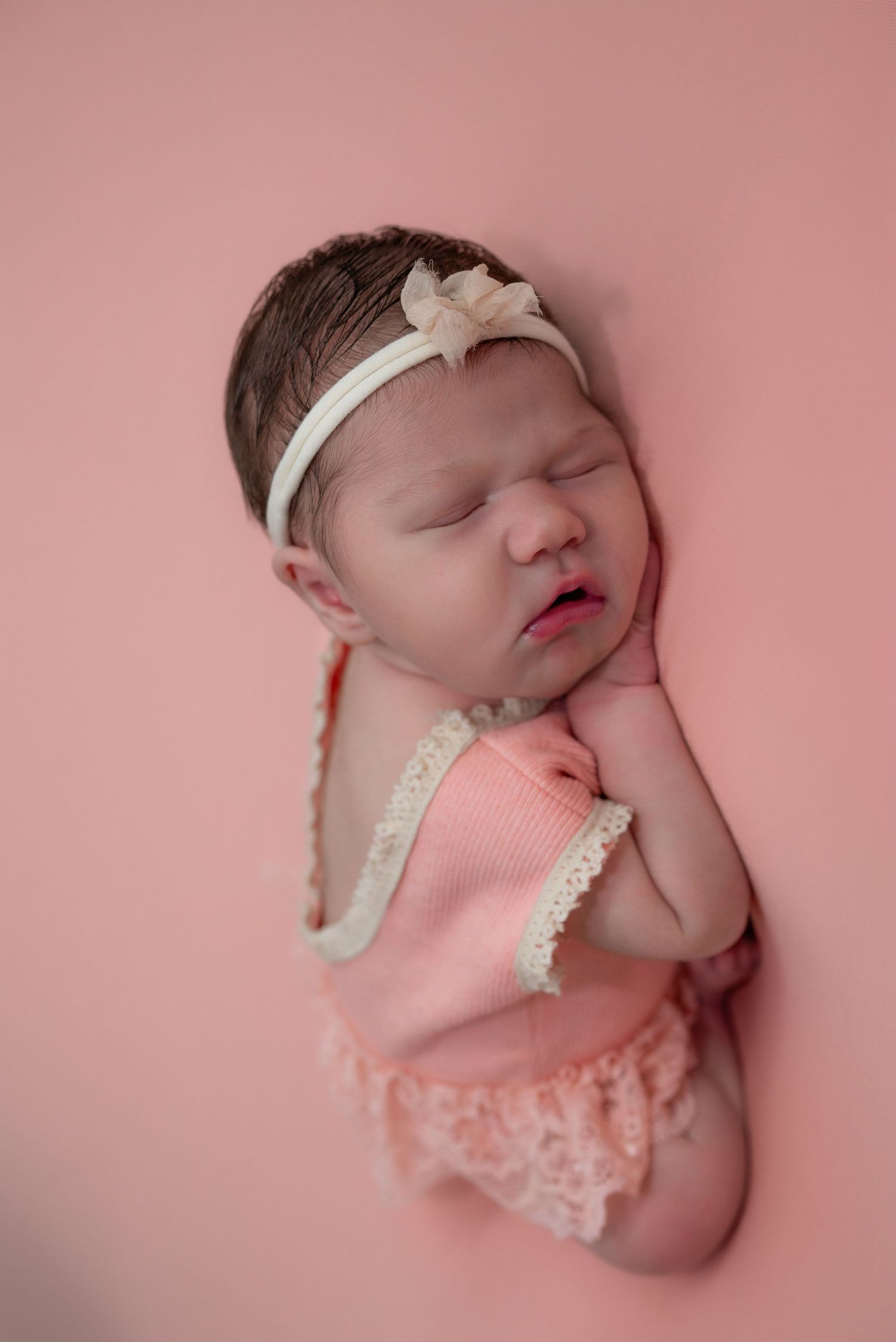Sleeping newborn baby girl in pink romper and bow headband posed on a pink background.