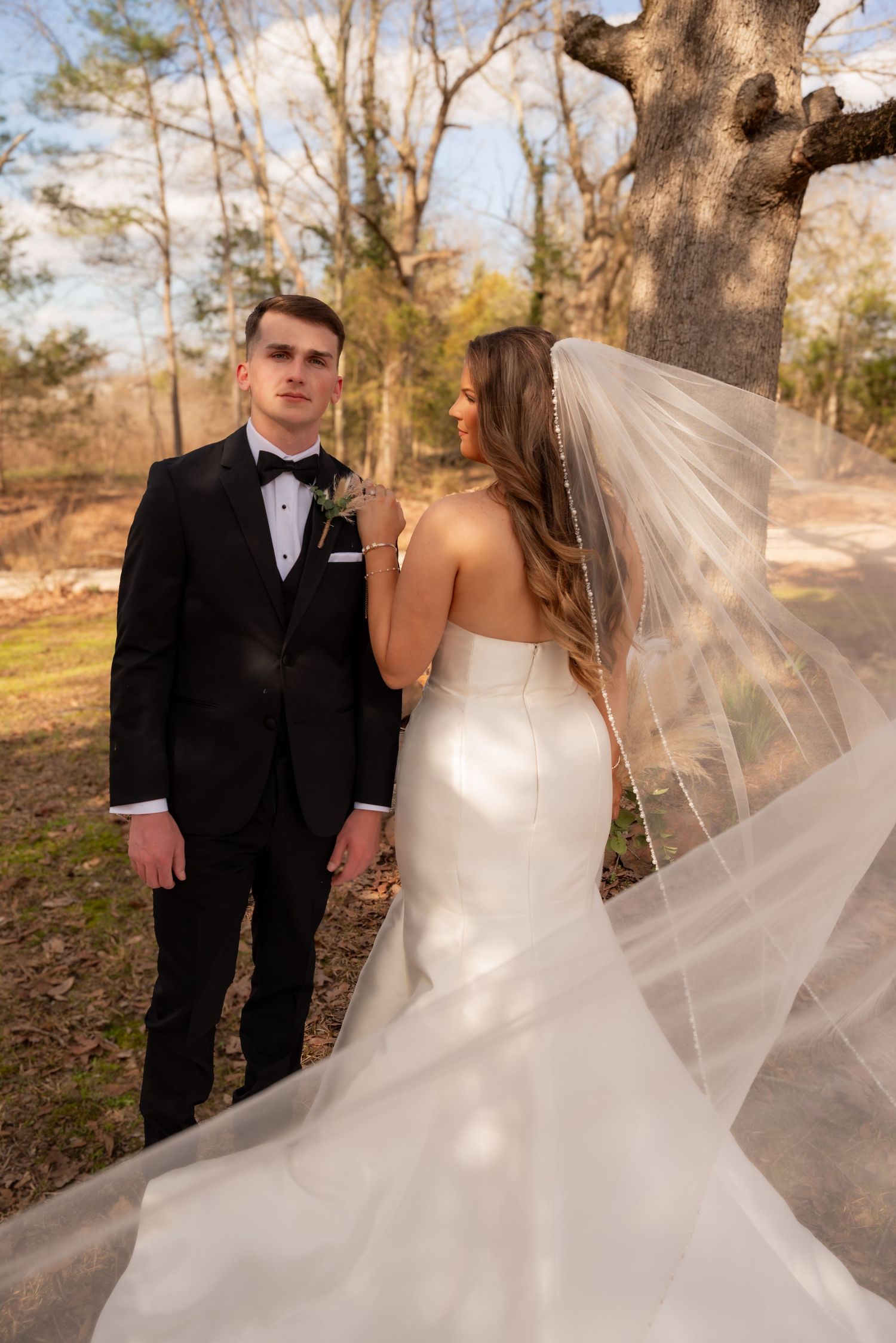 Bride in white gown with flowing veil stands beside groom in black tuxedo outdoors among autumn trees.