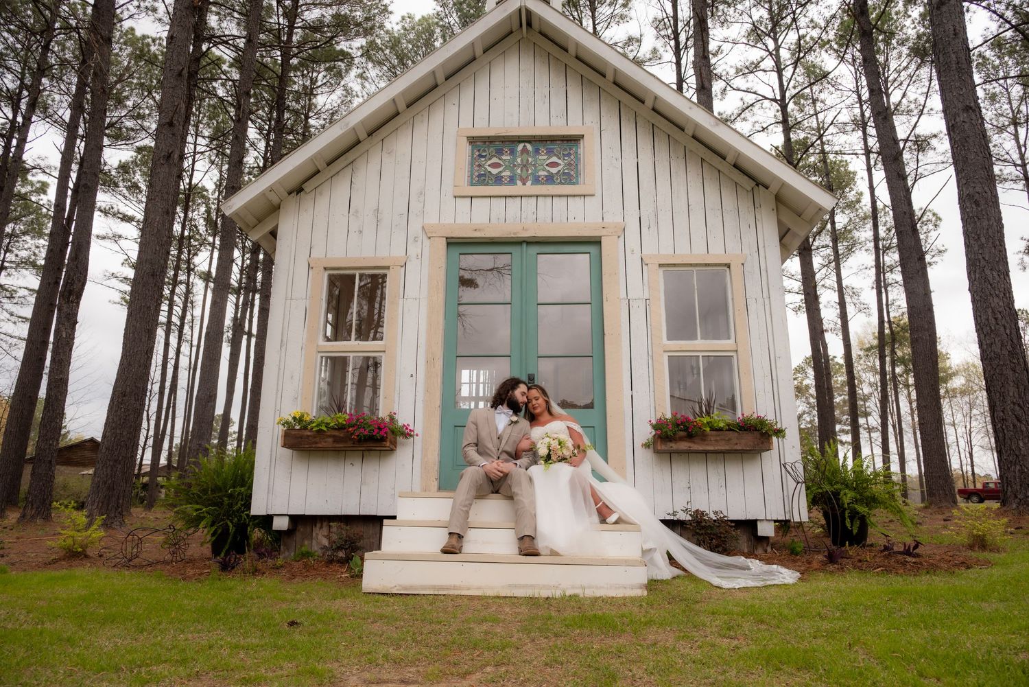 Bride and groom sitting on steps of charming white cottage surrounded by tall pine trees.