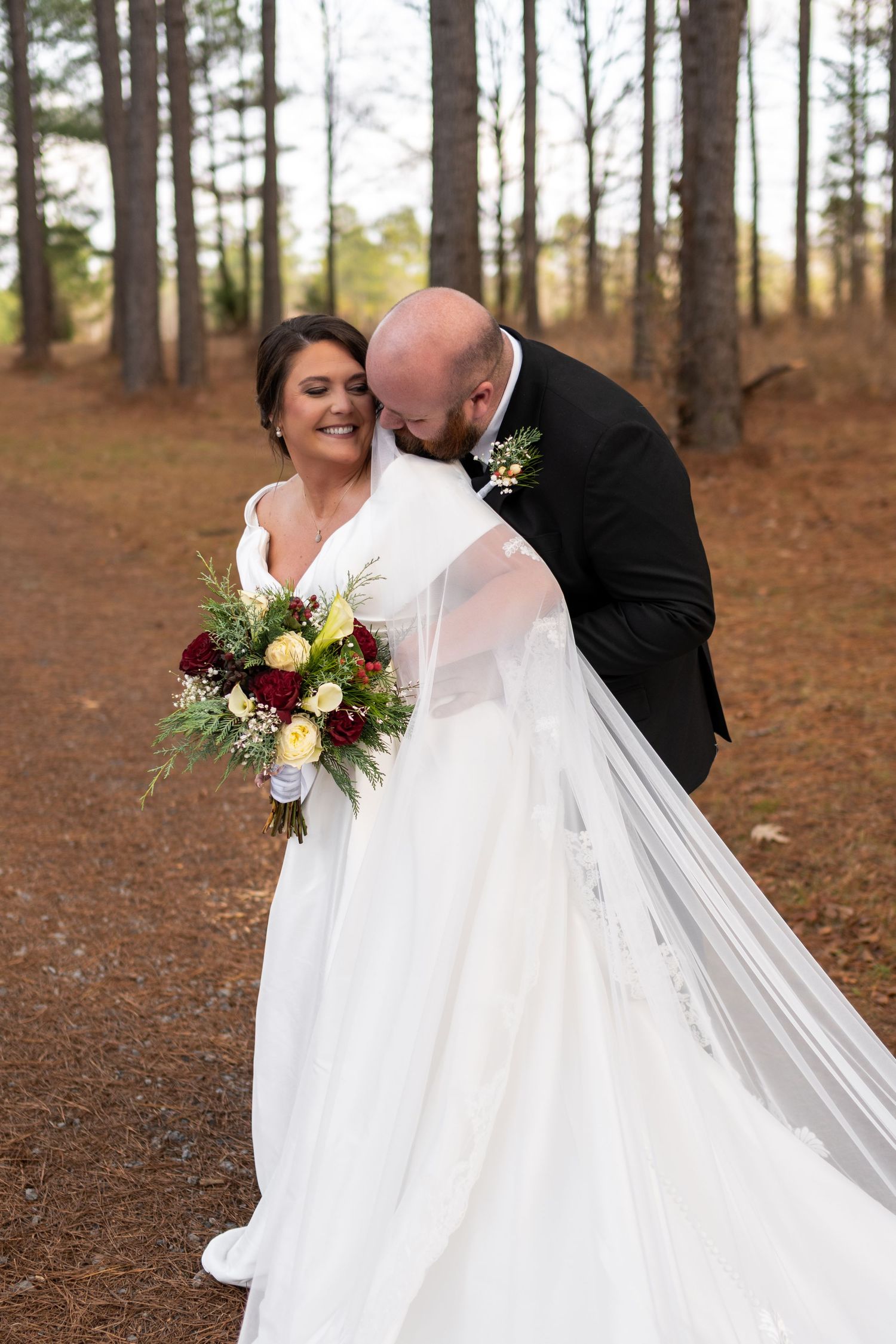 Bald groom in black suit kissing bride's cheek as she smiles holding red and white bouquet.