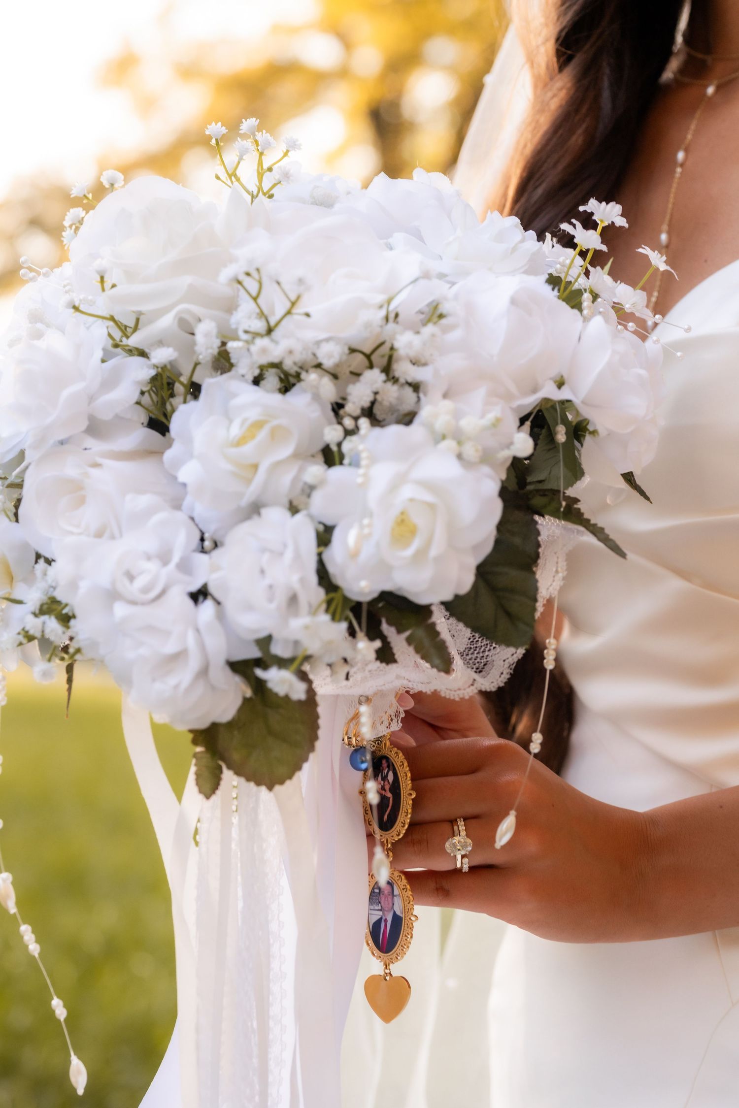 Bride holds a stunning white rose bouquet with gold charm accents, wearing a white wedding dress and diamond ring.