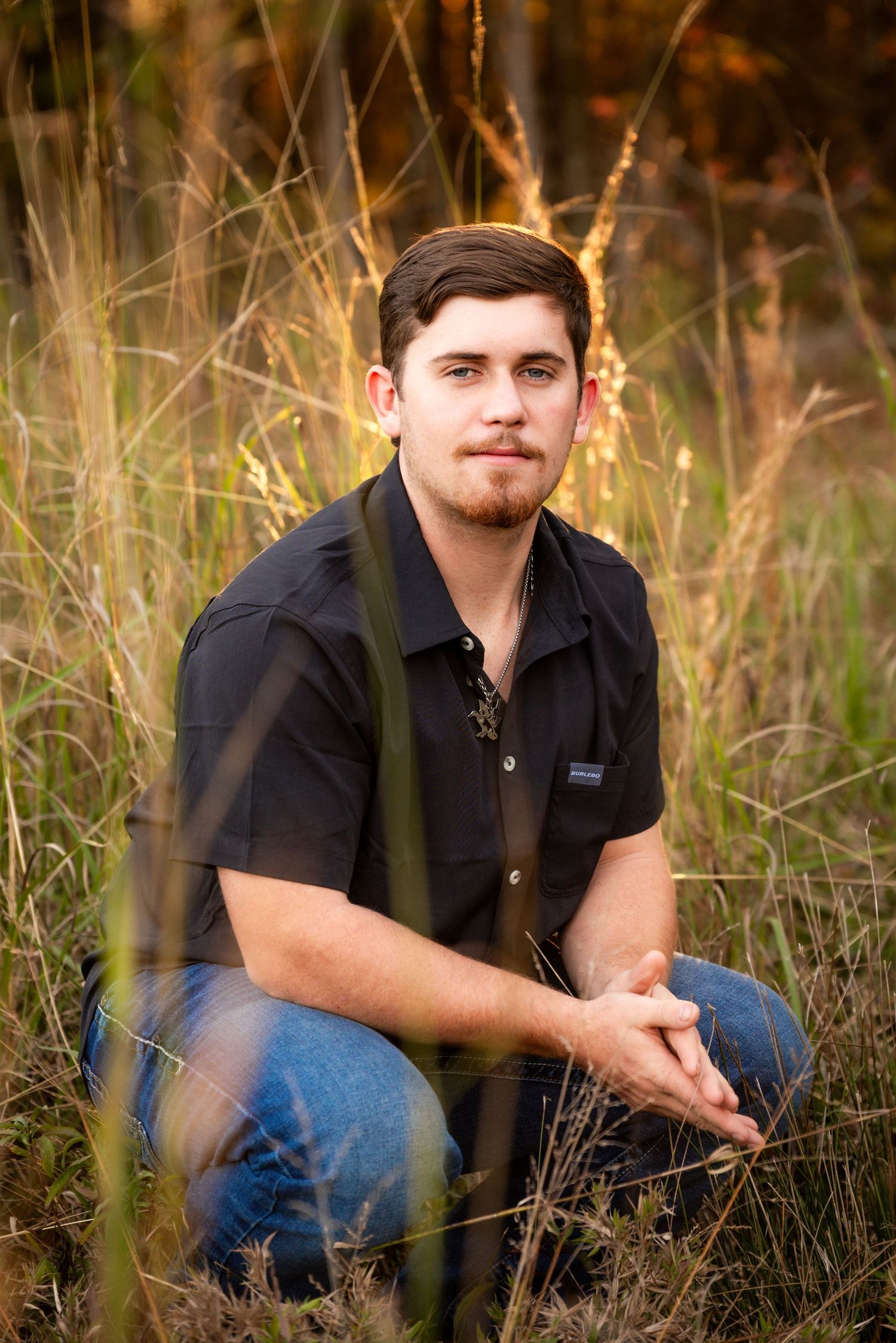 Young man in black shirt sitting in tall golden grass field during autumn sunset.