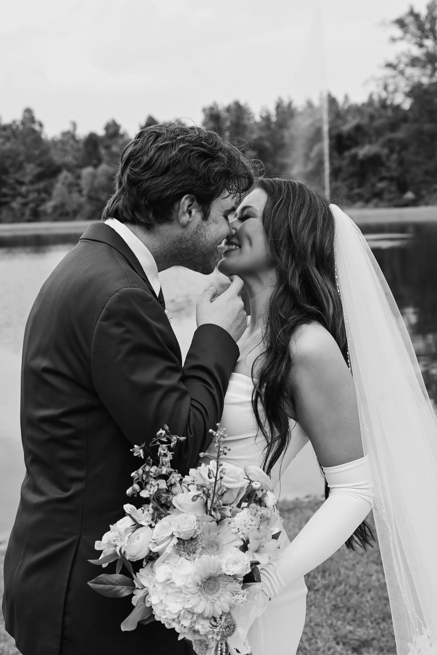 Black and white romantic photo of bride and groom nearly kissing by a lake, bride in long veil.