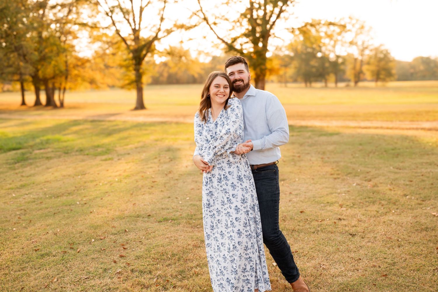 Smiling couple embracing in an open field during golden hour with autumn trees in background.