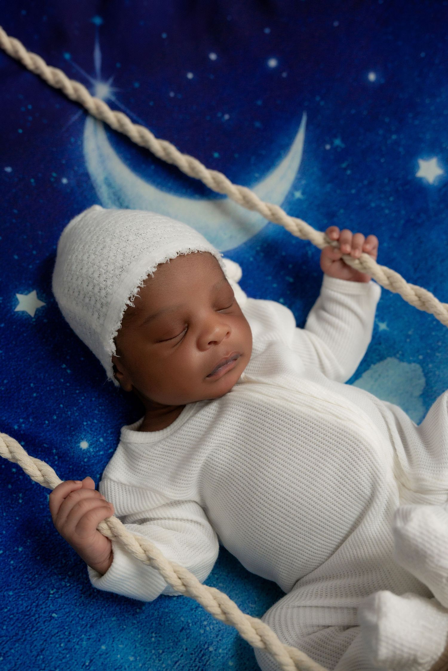 Newborn baby in white outfit and hat sleeping on a blue starry night themed backdrop.