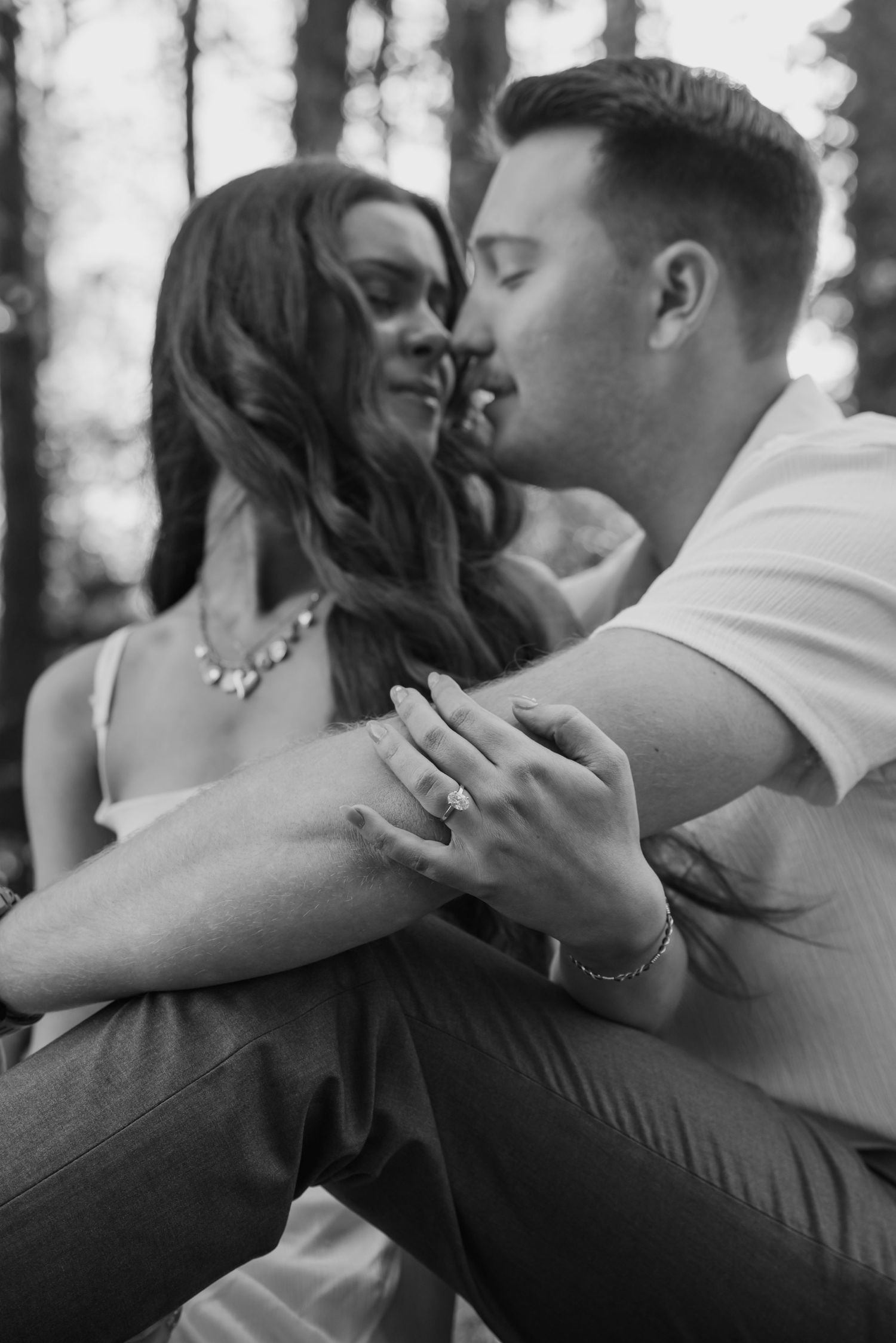 Black and white photo of couple embracing in forest, showcasing engagement ring on bride's hand.