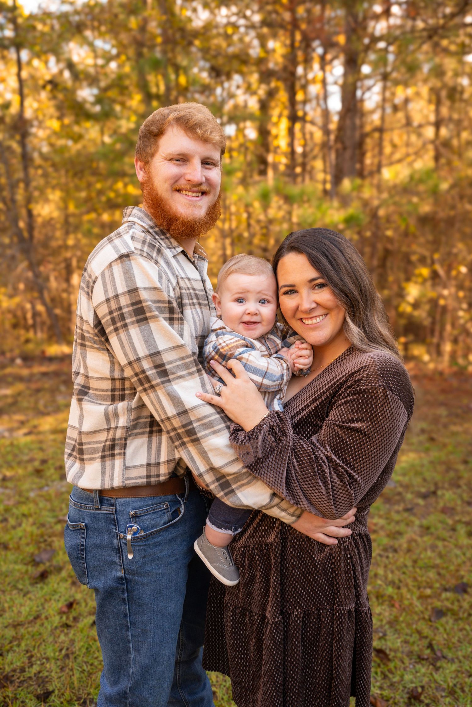 Young family with red-haired dad, brunette mom, and baby boy posing outdoors among fall foliage.