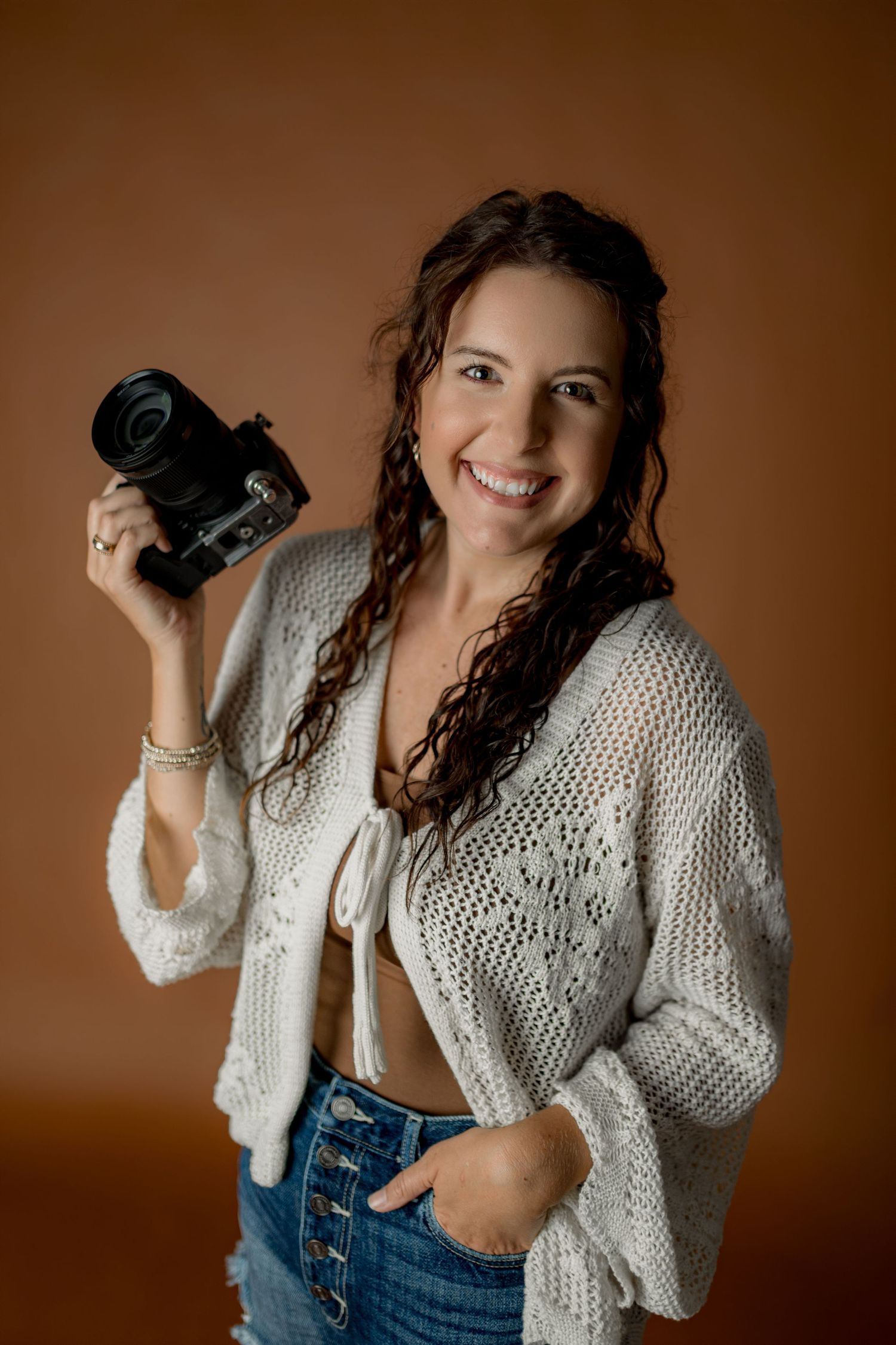 Smiling woman with curly hair holding a camera, wearing a white knit cardigan and jeans against a brown background.