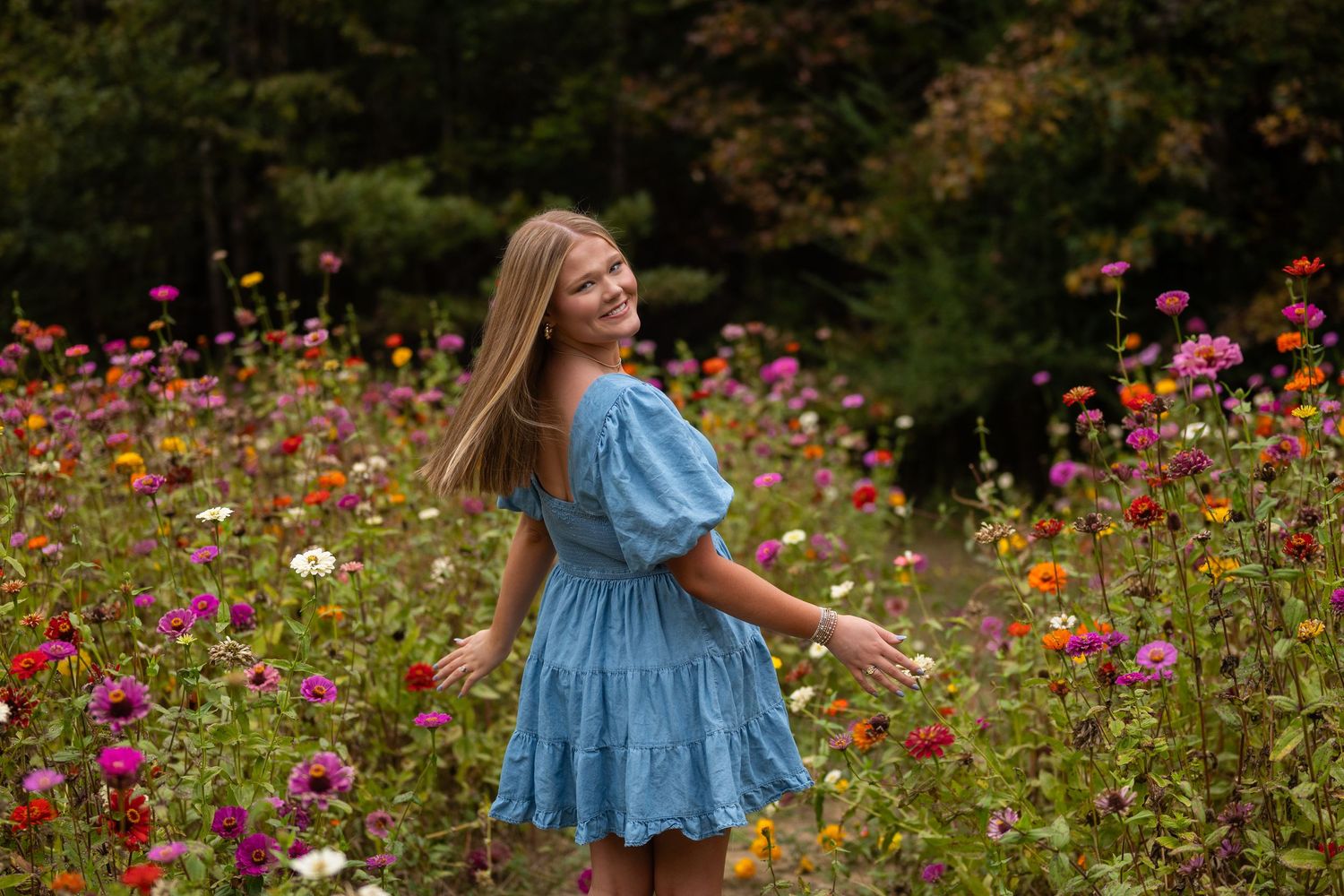 Blonde teenage girl in blue dress laughs joyfully surrounded by colorful blooming flowers outdoors.