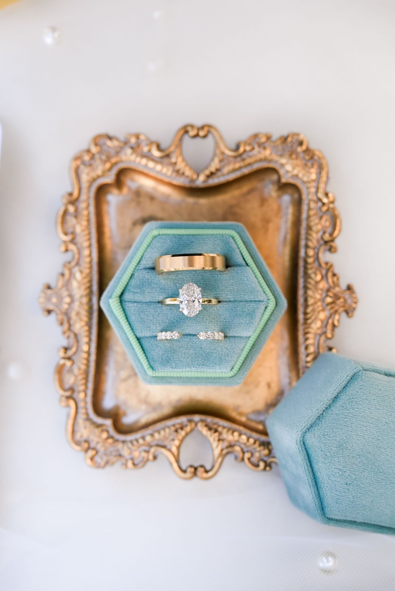 Oval diamond engagement ring and gold wedding band displayed in teal velvet hexagon ring box on ornate gold tray.