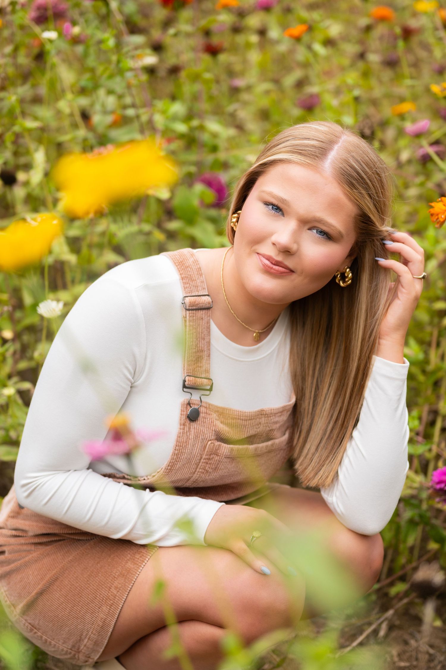 Teen girl in brown overalls sitting among colorful wildflowers for senior portrait.