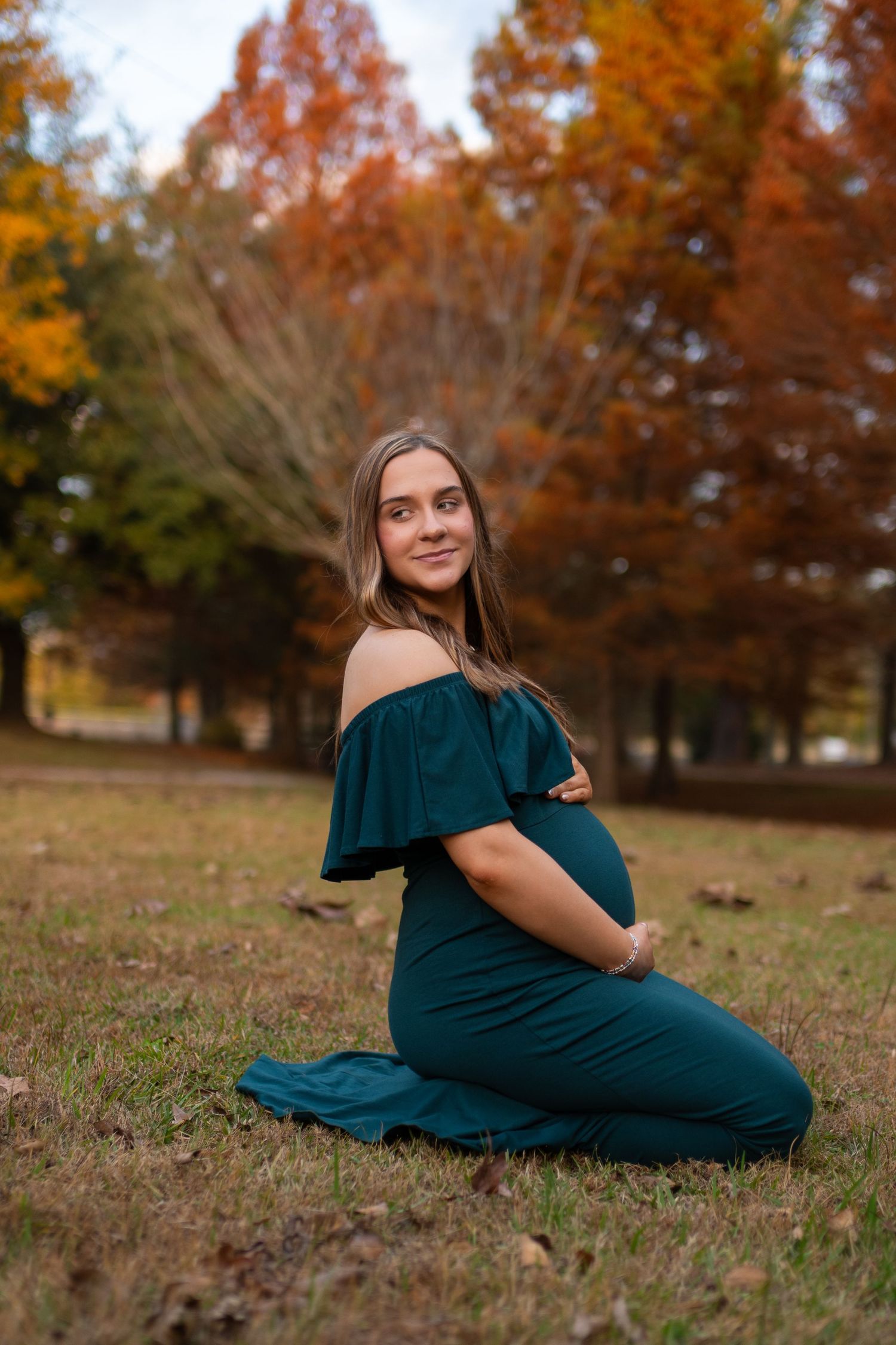 Pregnant woman in teal off-shoulder gown sitting outdoors among fall foliage trees.