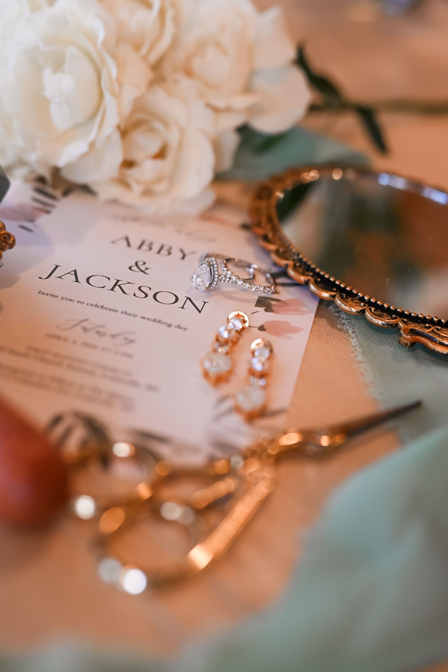 Wedding invitation for Abby and Jackson surrounded by pearl earrings, roses, and gold scissors.