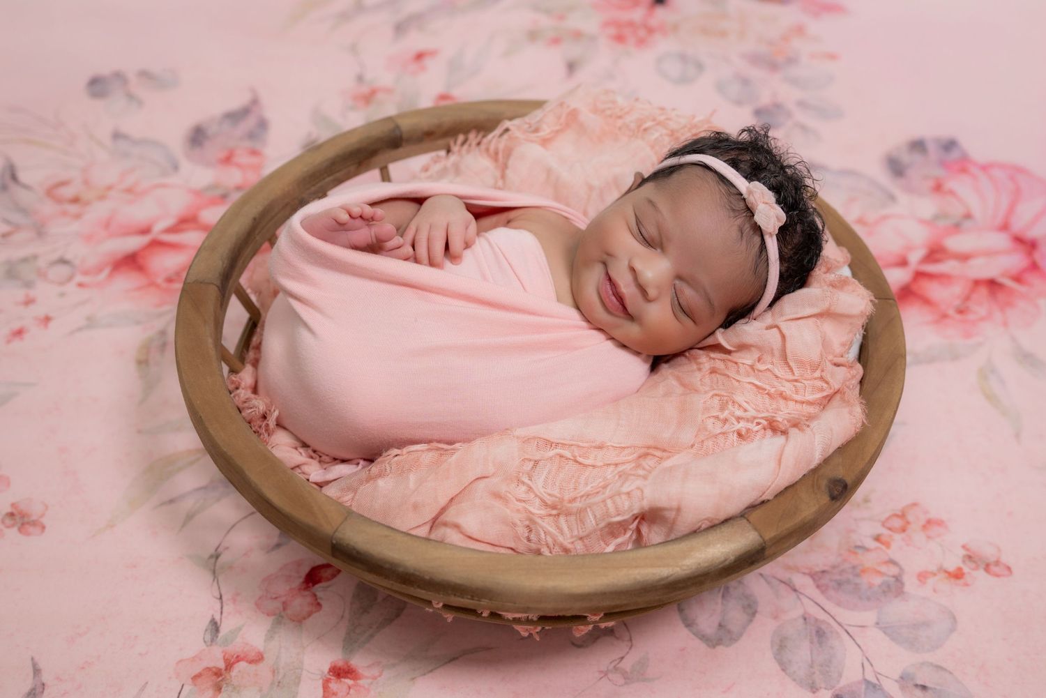 Smiling newborn baby girl wrapped in pink swaddle with lace headband sleeping in wooden bowl prop.